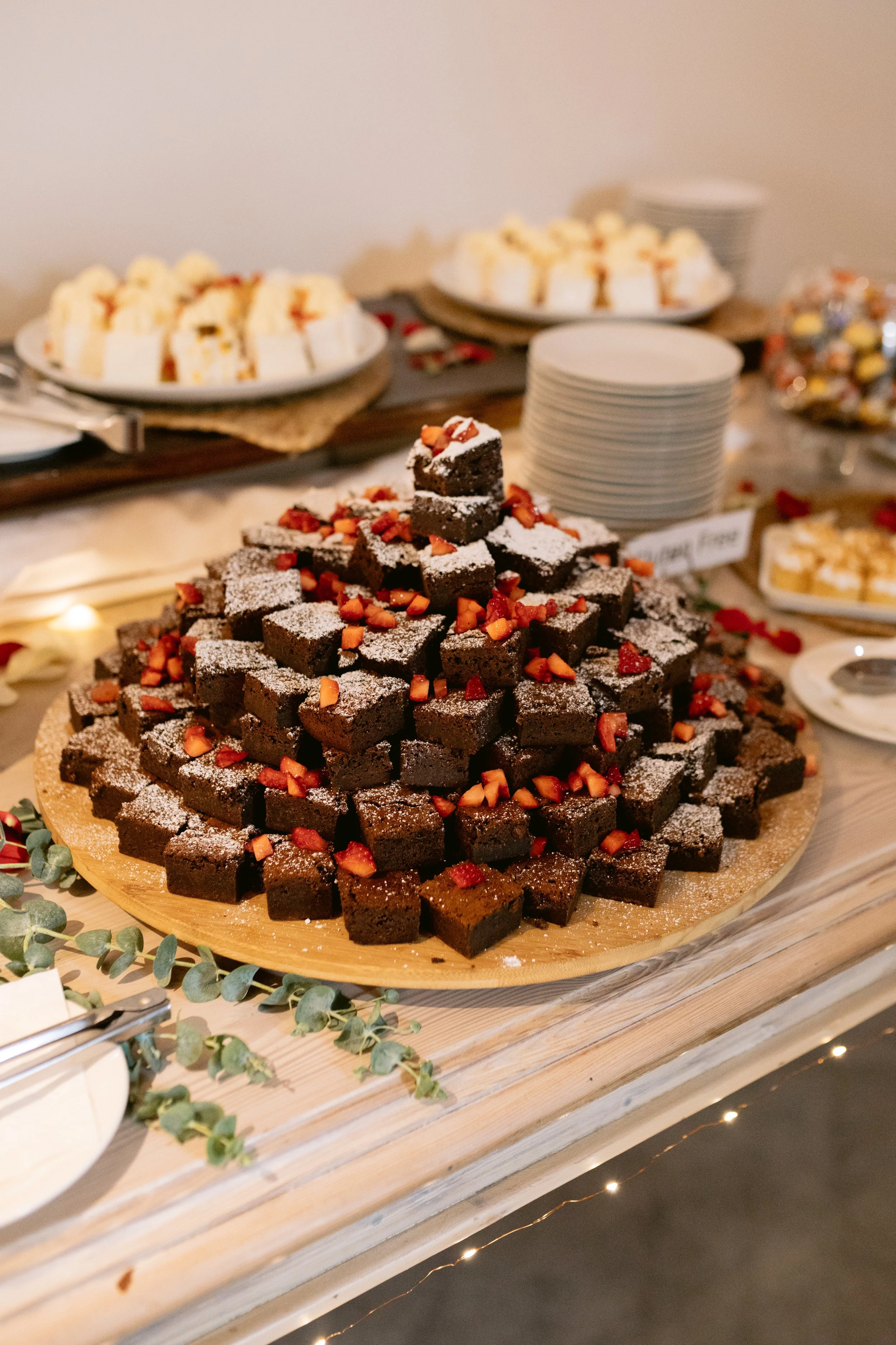 Chocolate brownies topped with powdered sugar and small pieces of strawberries, arranged in a pyramid on a wooden serving board at a dessert table.