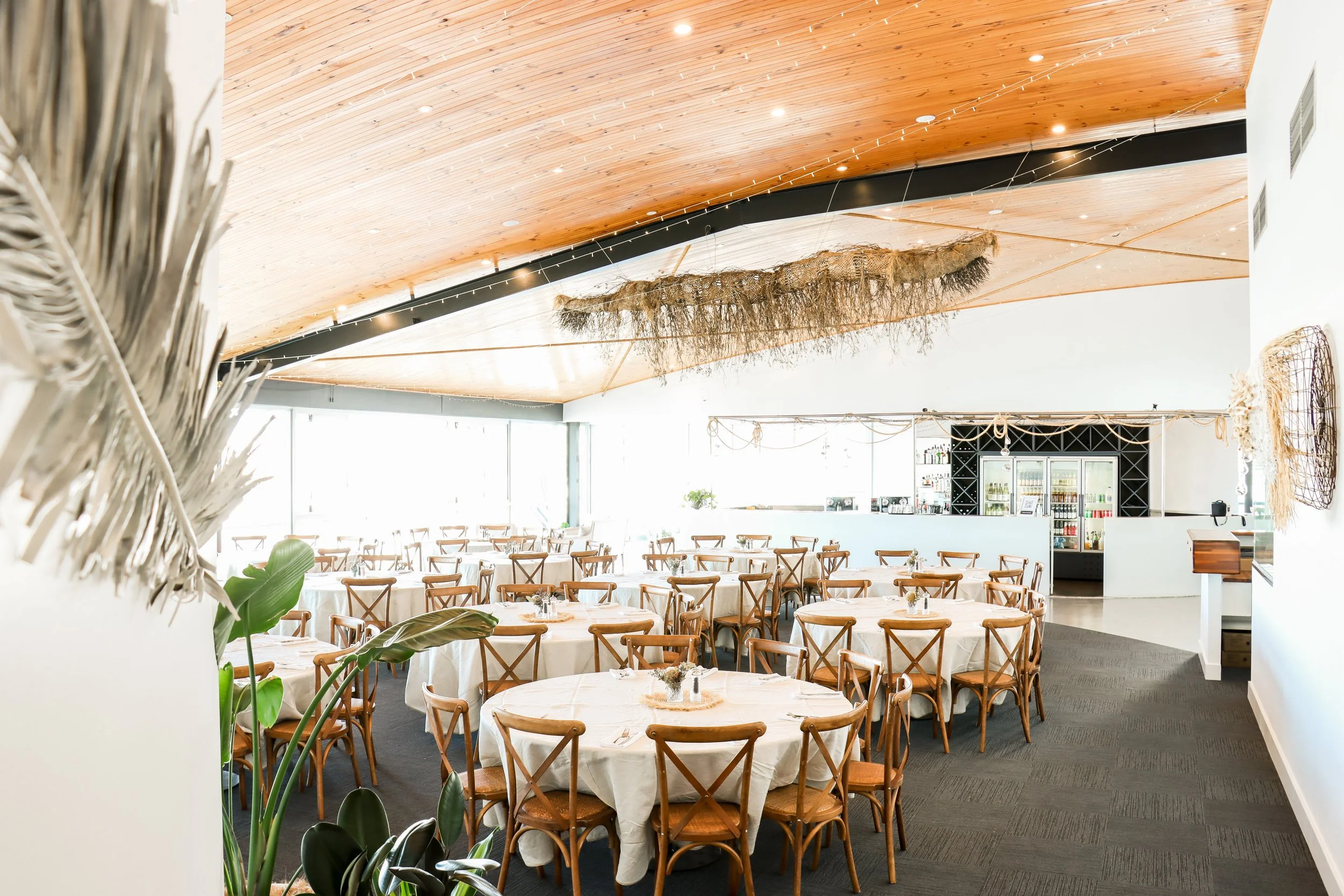 Interior of a bright, modern restaurant with round tables covered with white tablecloths, surrounded by wooden chairs. The ceiling has a curved wooden design with string lights hanging. Large windows at the back let in natural light, and there is a bar area in the background with shelves of beverages.