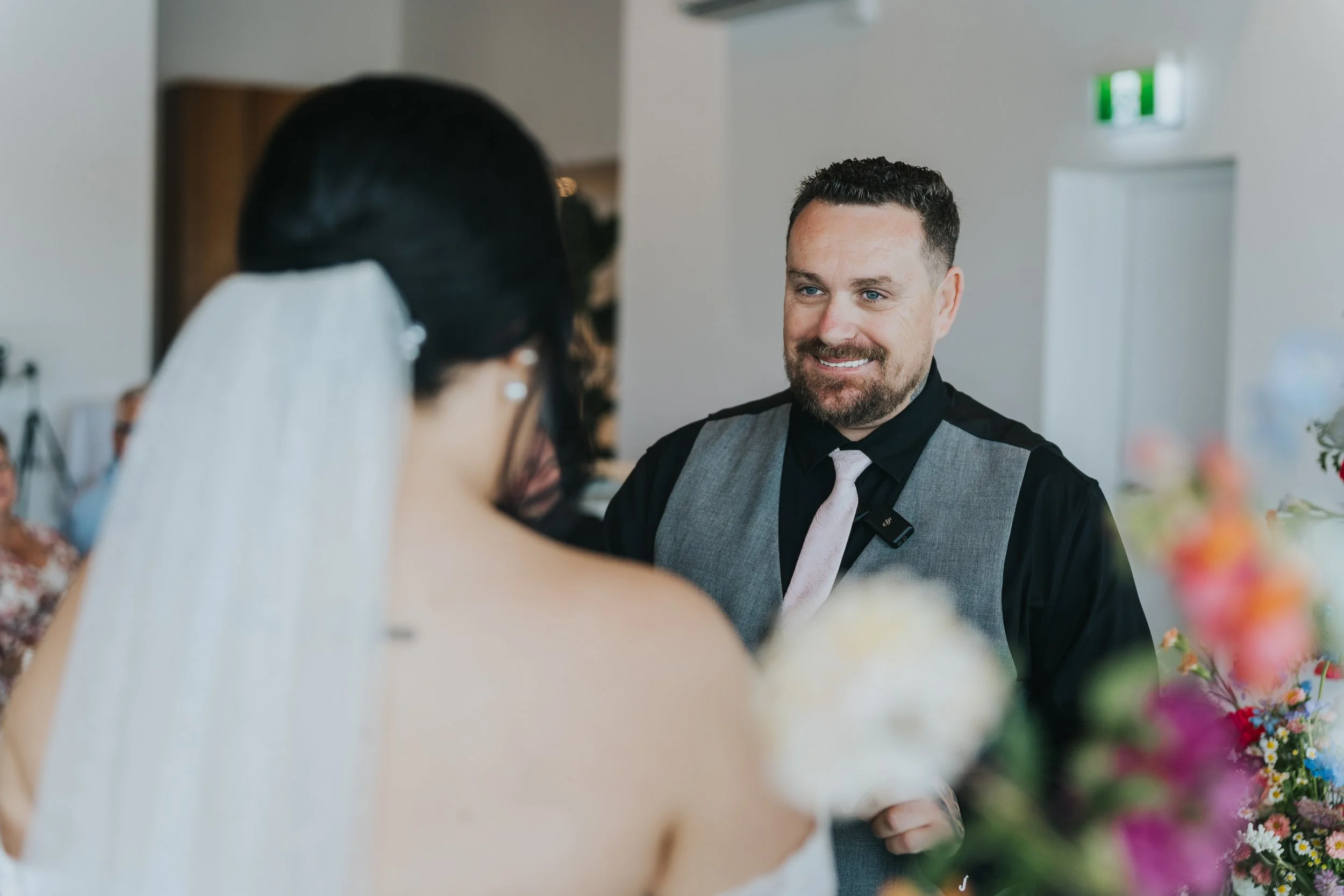 A groom with a beard and a black shirt, gray vest, and light tie, smiling at a bride with dark hair, veil, and earrings, during a wedding ceremony indoors.