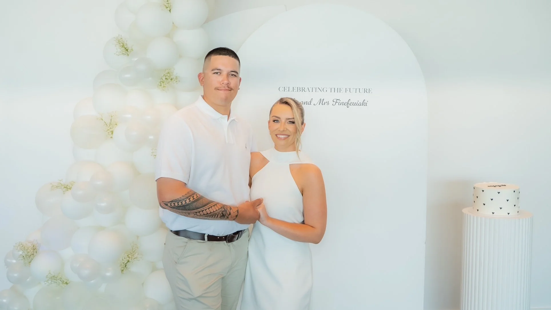 A newlywed couple holding hands and smiling at a celebration event with white balloons and a tiered cake.