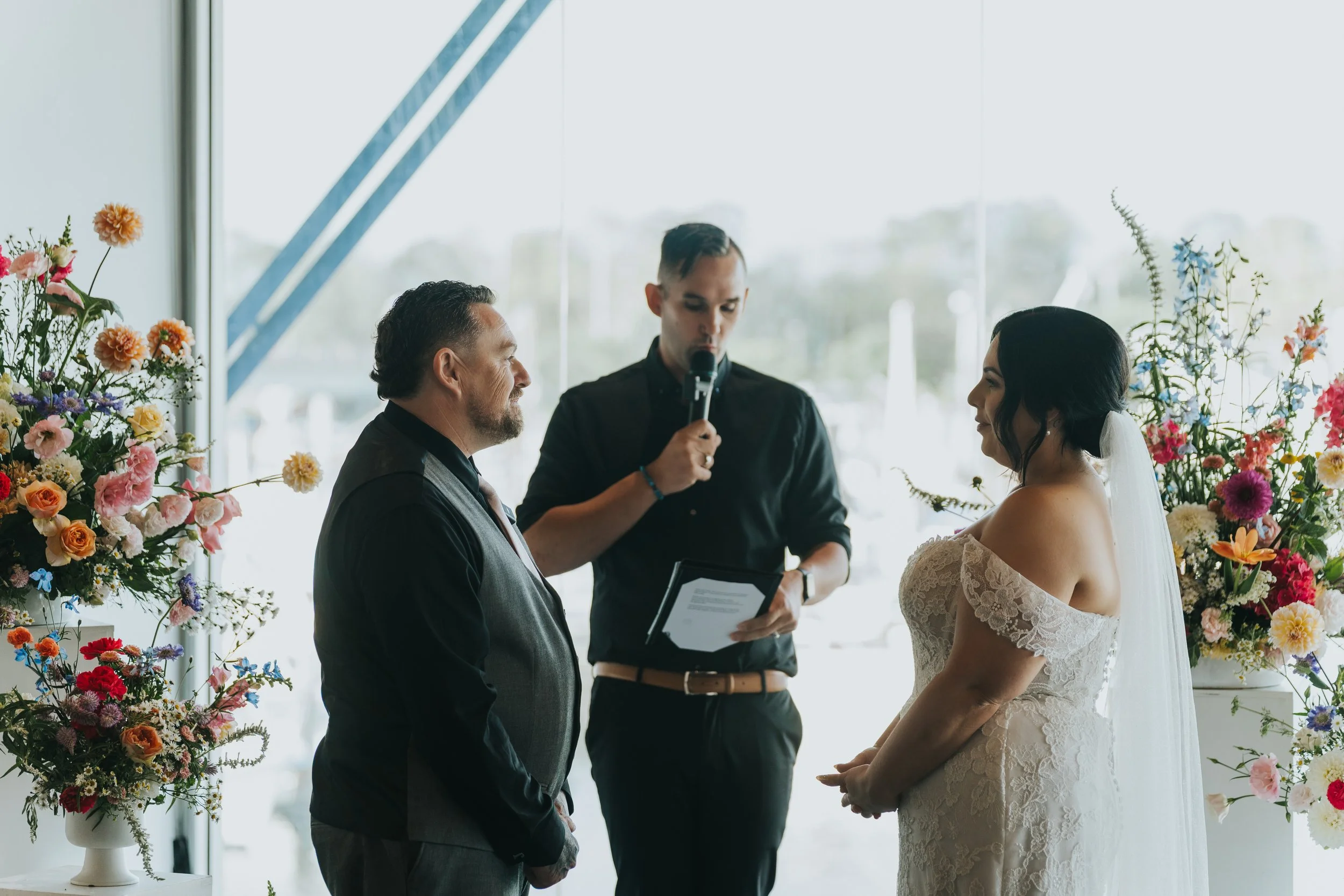 A couple getting married, standing face-to-face with an officiant, during a wedding ceremony inside a venue decorated with colorful flowers.