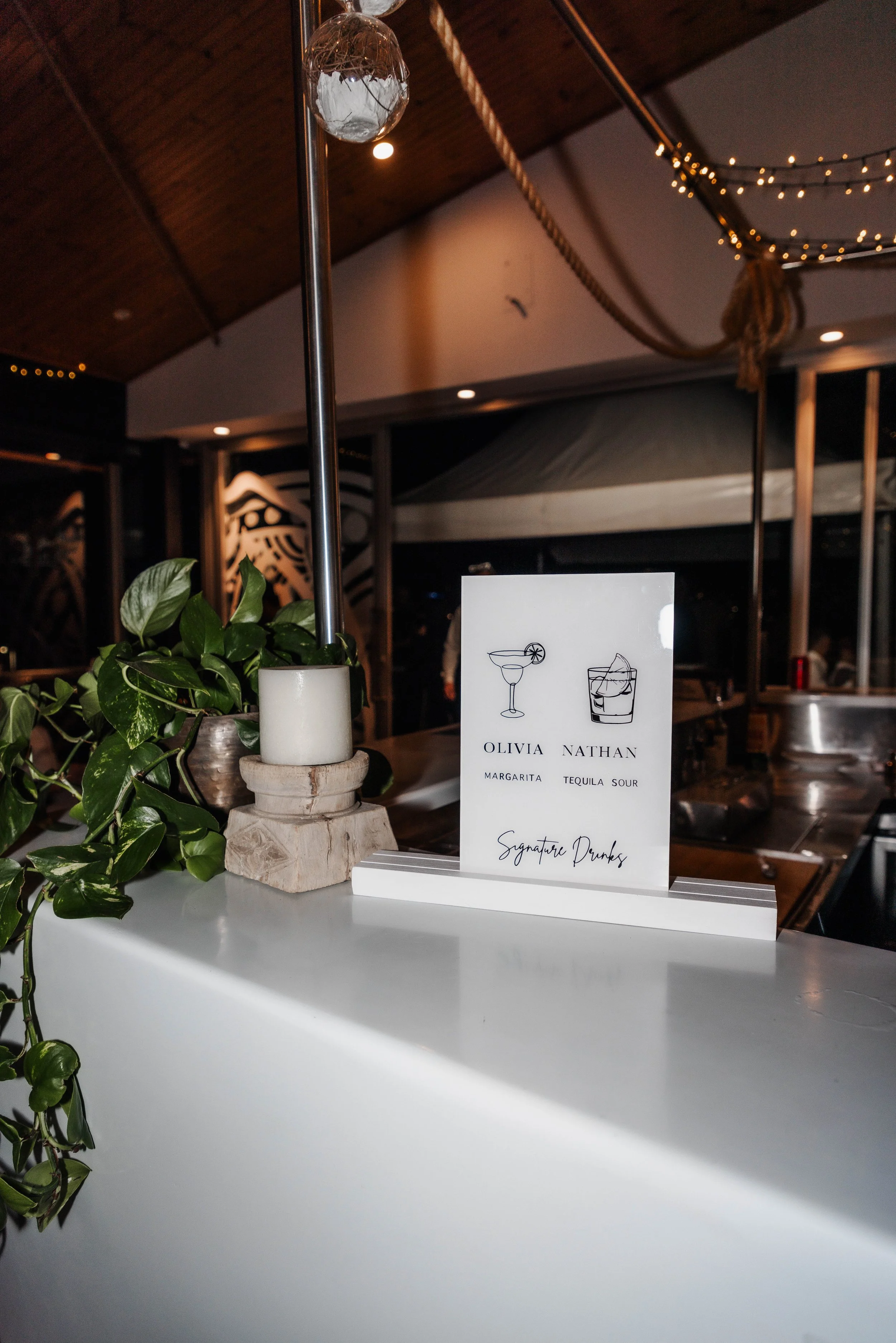 A table set at a bar with a black sign displaying the names Olivia Nathan, with illustrations of a margarita and a tequila sour, and the text 'Signature Drinks.'