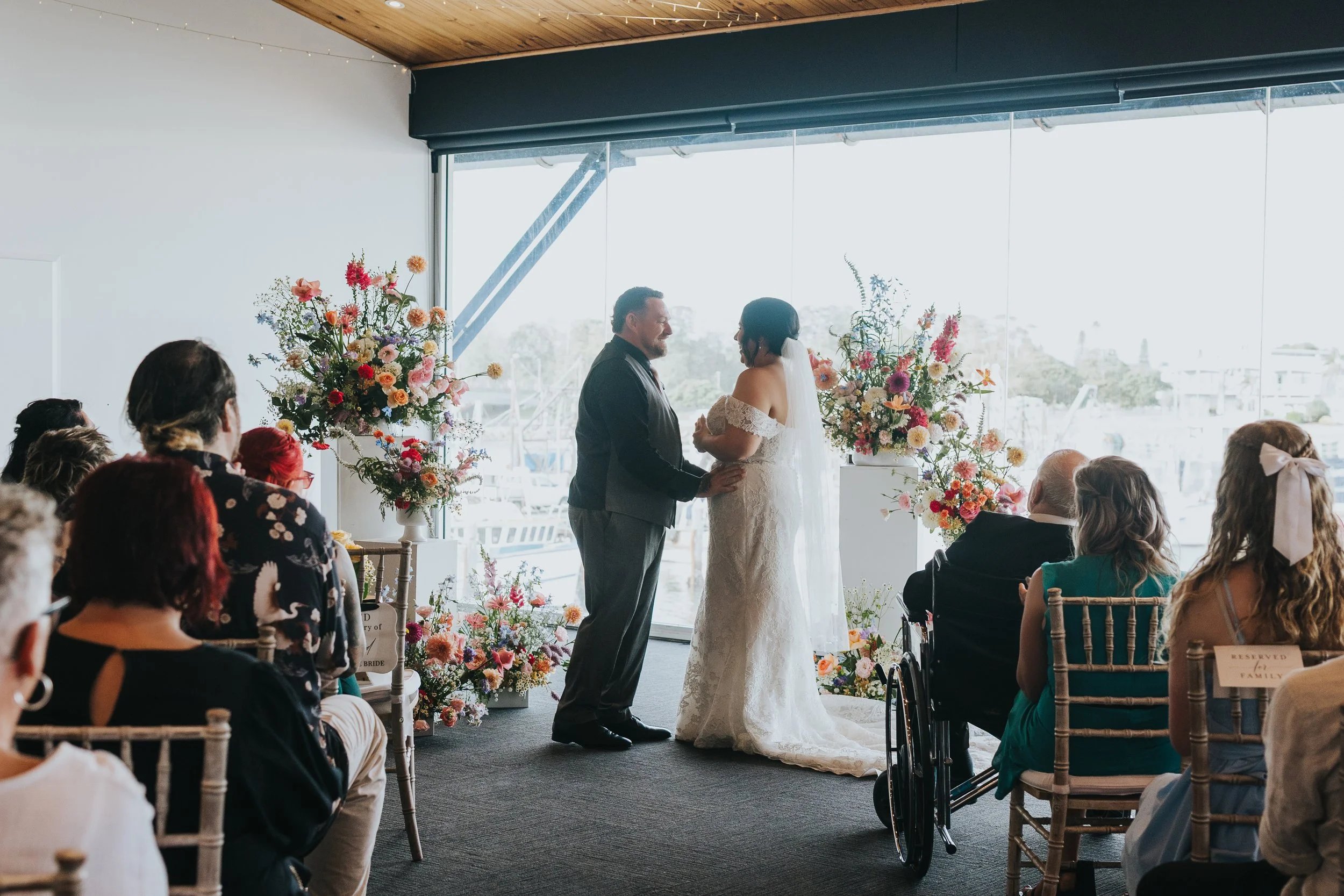 A wedding ceremony with a bride and groom standing in front of a large window, exchanging vows, surrounded by large floral arrangements. Guests seated on chairs are observing the event, some in wheelchairs, in a modern venue overlooking a marina.