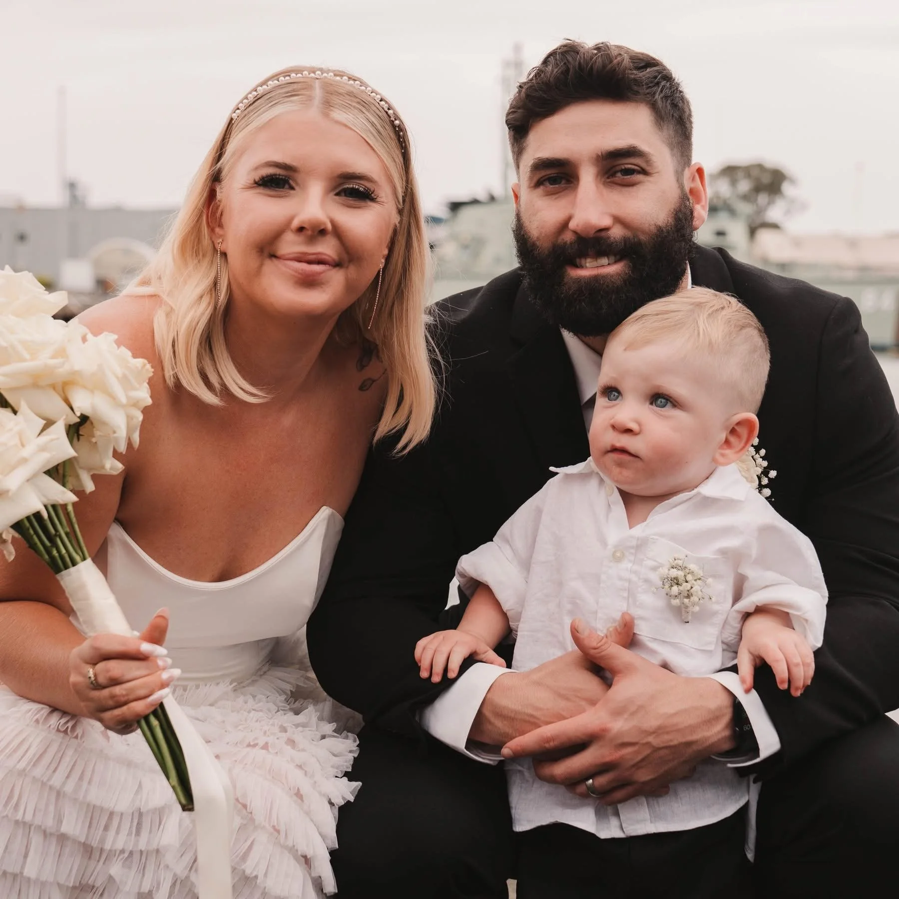 A family celebrating a wedding outdoors. A woman in a white dress and pearl headband, a man in a black suit, and a young boy in a white shirt, sitting together with a cityscape and boats in the background.