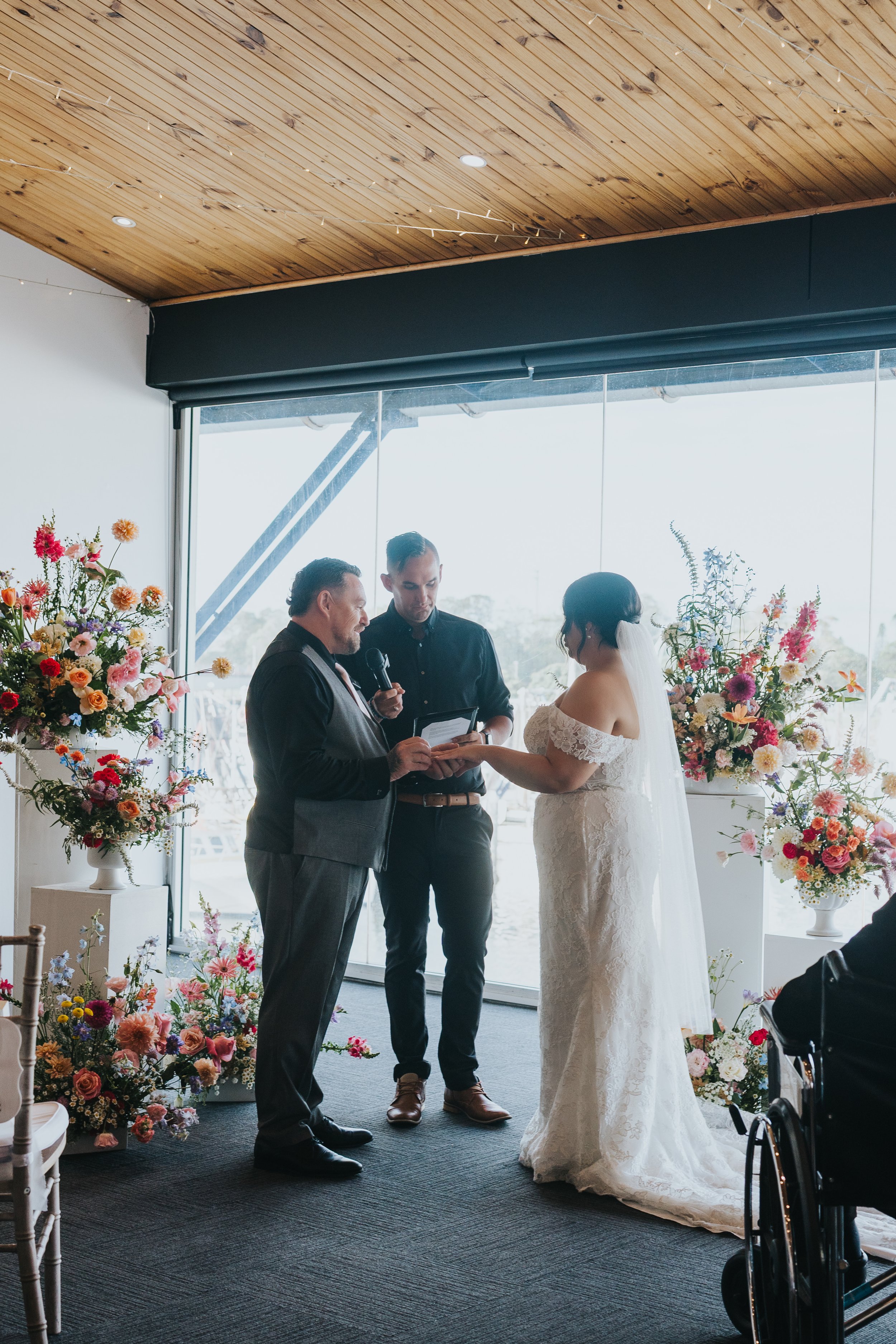 A bride and groom hold hands during their wedding ceremony with an officiant and an individual holding a microphone, surrounded by large floral arrangements in a bright indoor space.