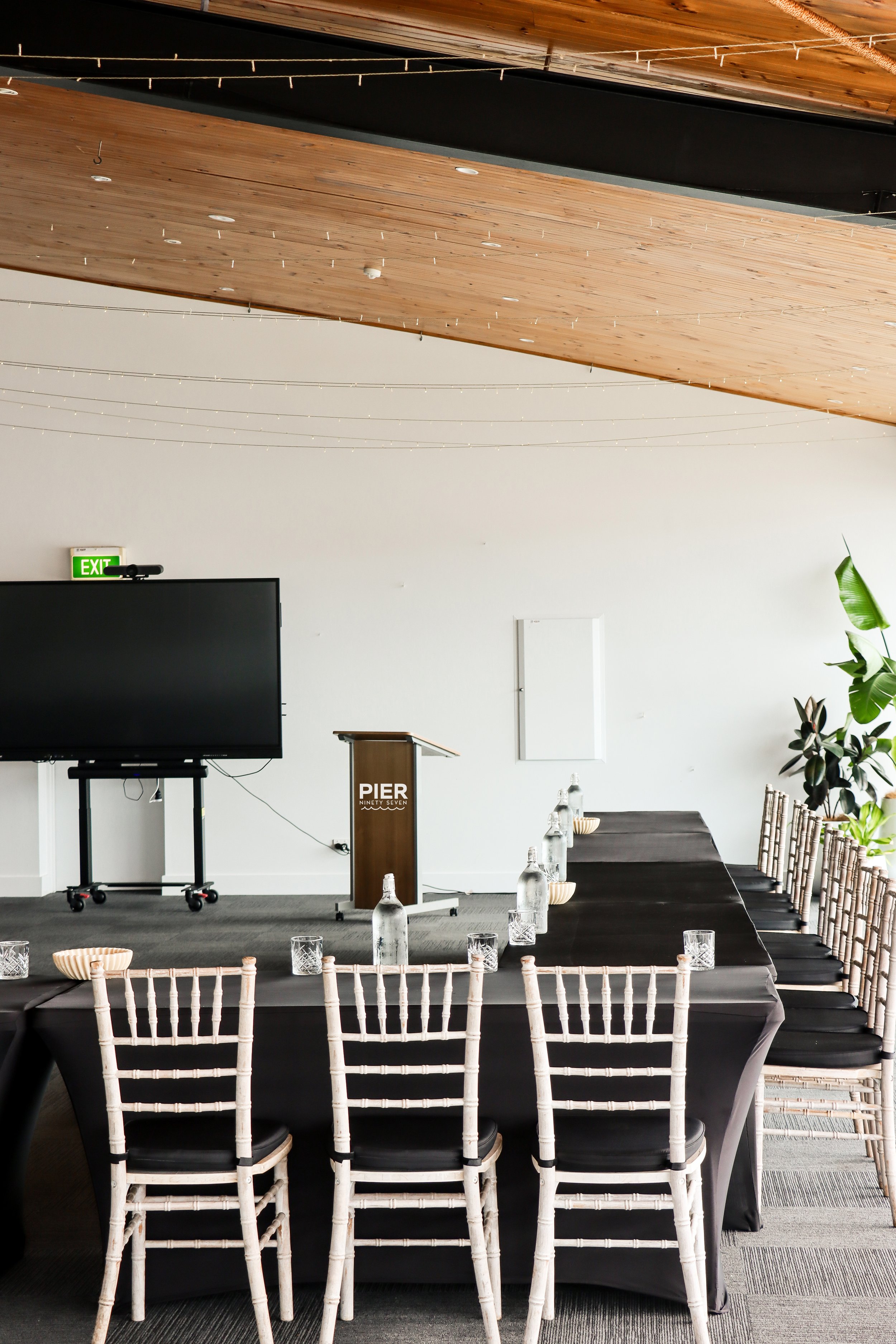 Empty conference room with a U-shaped table set with water bottles and glasses, a podium labeled "Pier," a large TV monitor, a whiteboard, and seating with white chairs with black seats, decorated with string lights and large green plants.