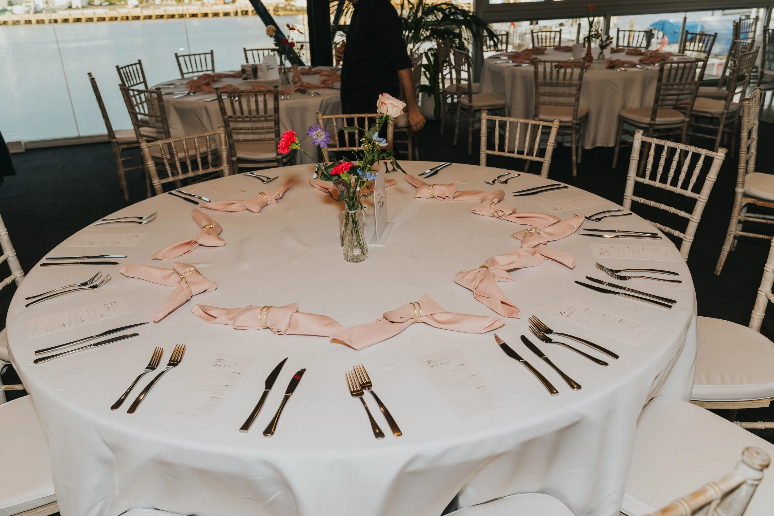 Round banquet table set for a formal event with a white tablecloth, pink napkins tied with rings, silverware, and a small floral centerpiece with pink and purple flowers, located near a window overlooking water.