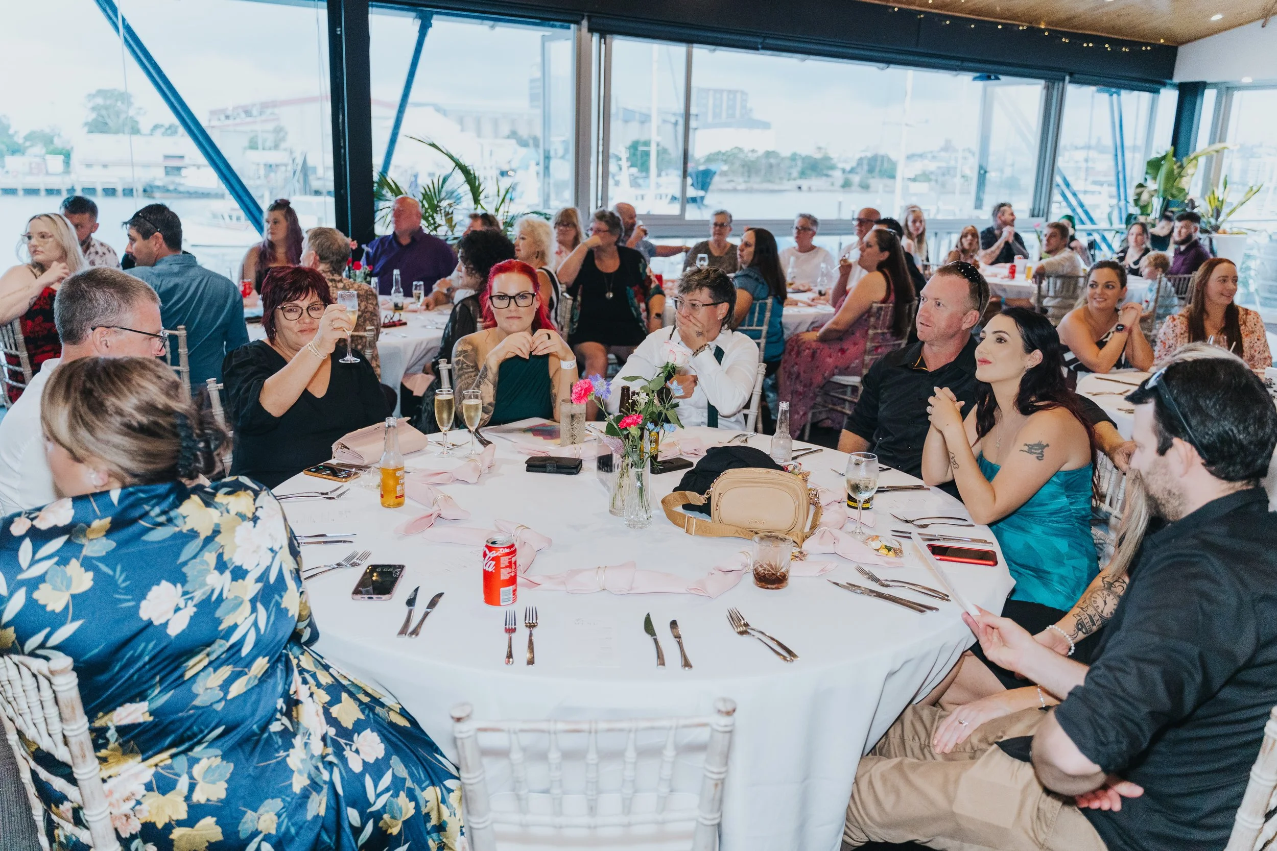A group of people seated at a round dining table during a social event or celebration at a venue with large windows and a view of boats and buildings outside.