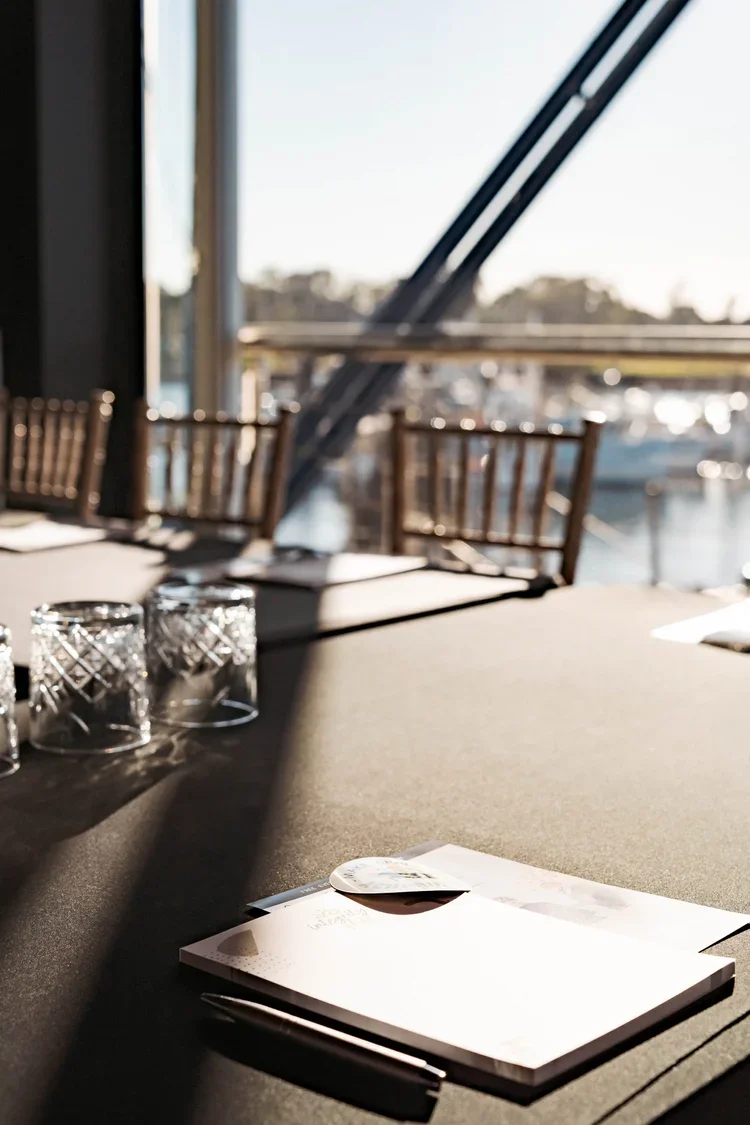 A restaurant or cafe table at a waterfront location with sunlight casting shadows. The table has glassware, a menu, and a pen. There are wooden chairs and a large window showing boats and a marina outside.