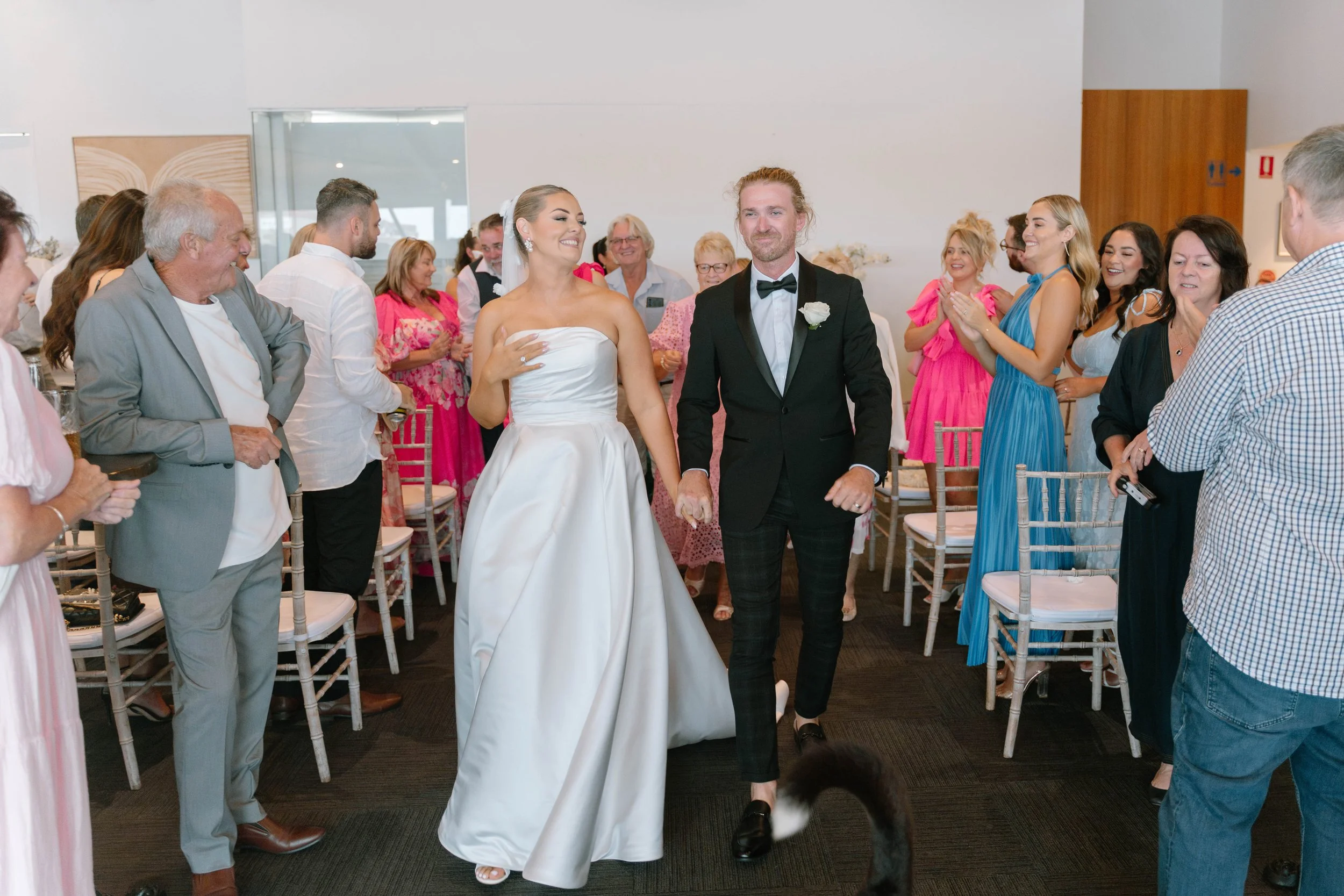 Bride and groom walking down aisle during wedding reception, surrounded by guests clapping and smiling.