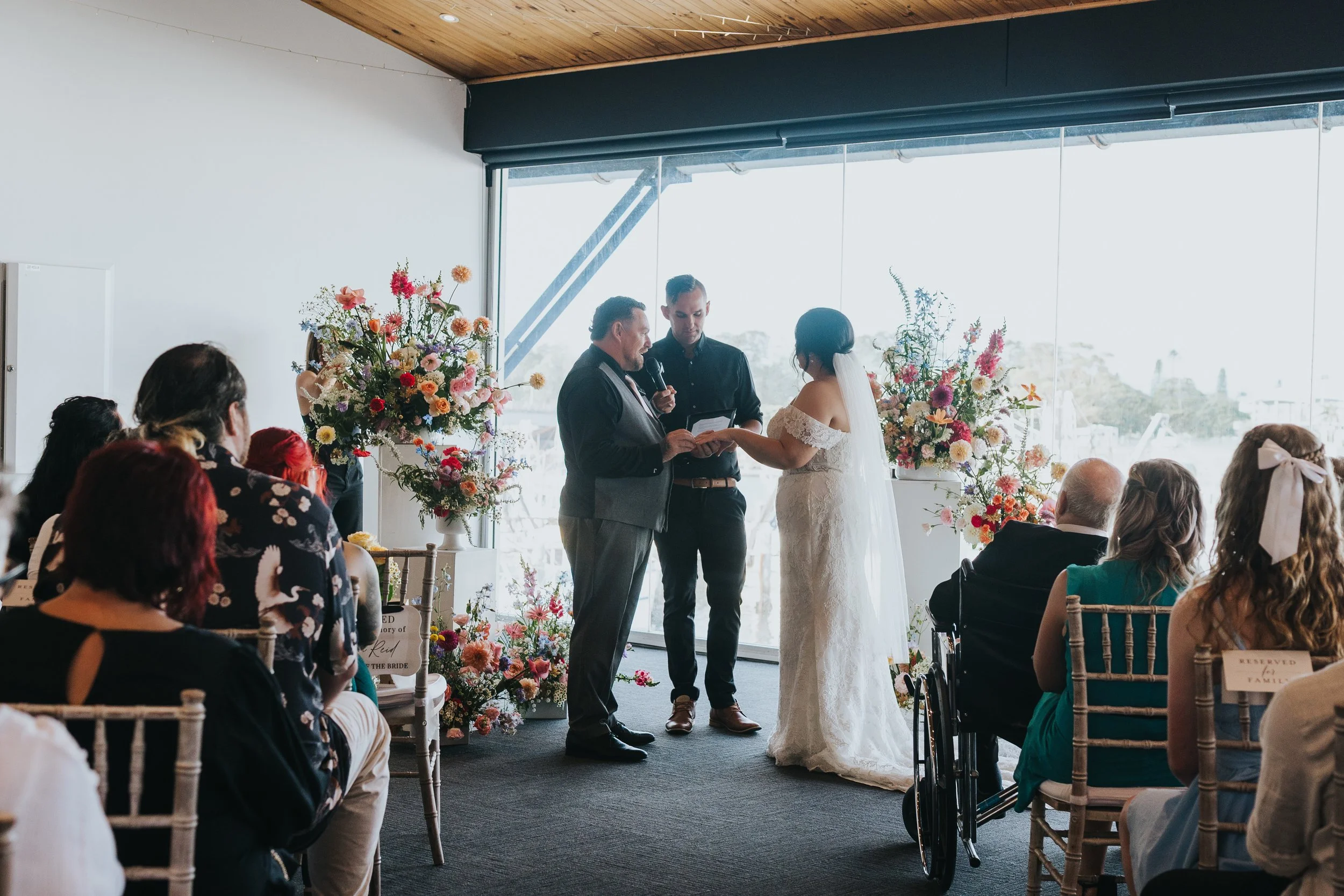 A wedding ceremony with a bride and groom exchanging vows in front of guests, surrounded by floral arrangements, with a large window in the background letting in natural light.