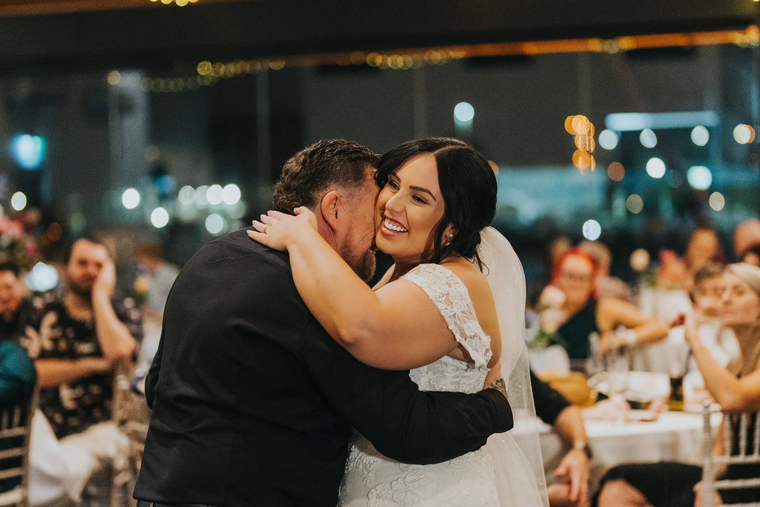 Bride and groom hugging and smiling at their wedding reception, with guests seated at tables in the background.