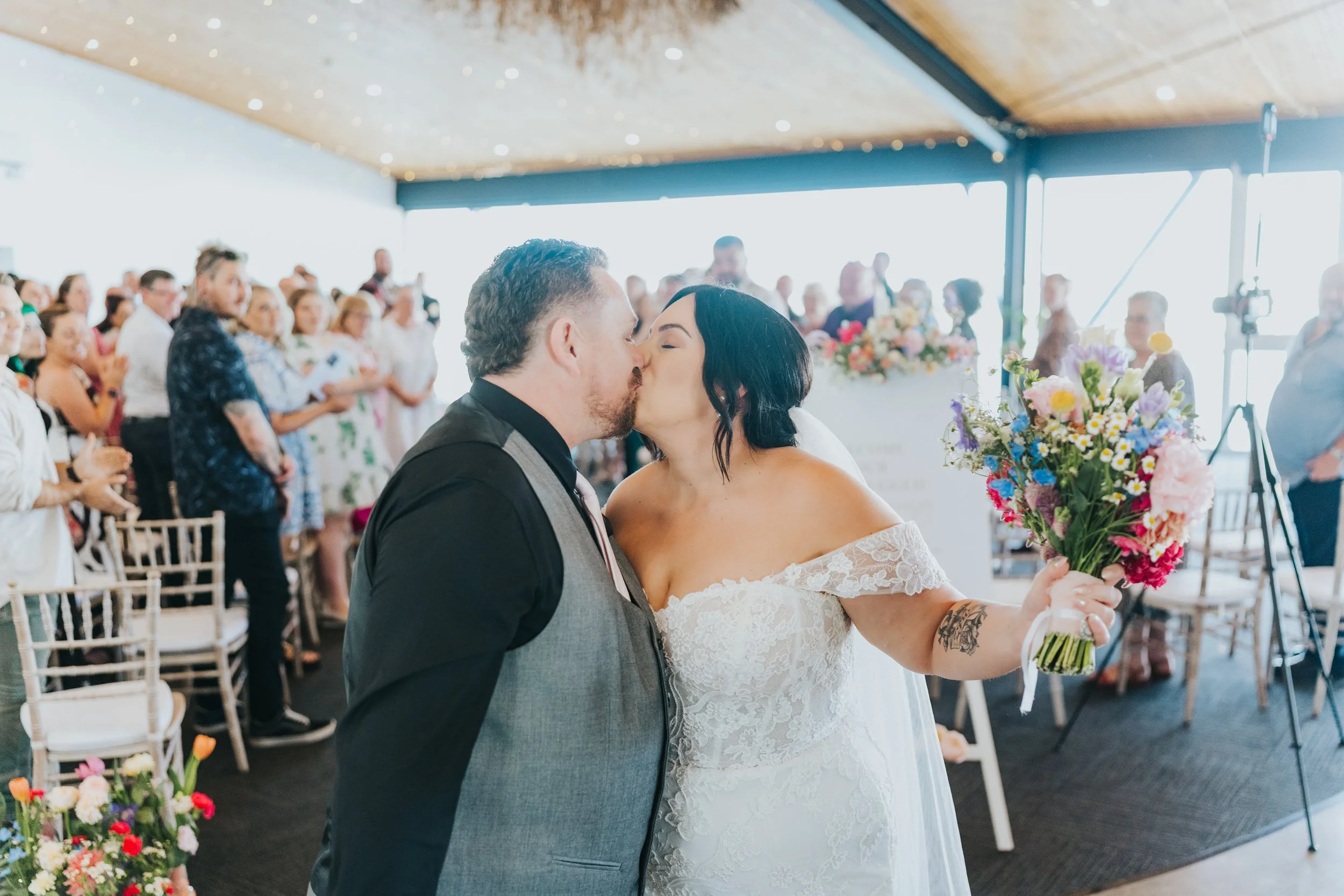 A bride and groom kissing during their wedding ceremony, with wedding guests in the background clapping and smiling.