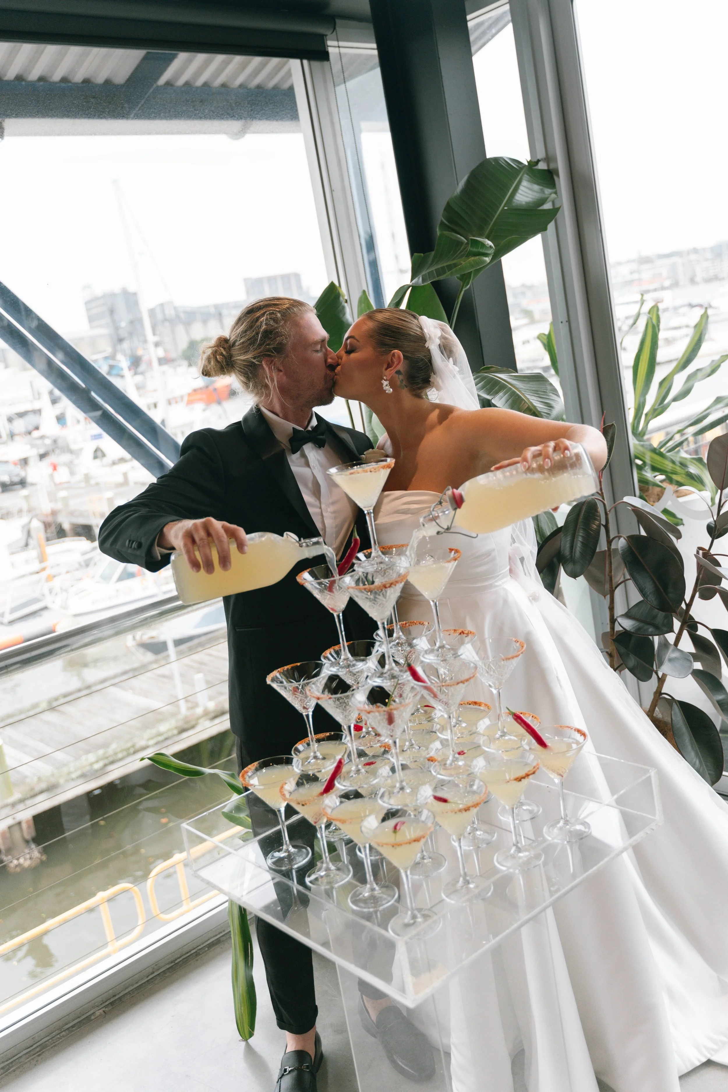 Bride and groom sharing a kiss during their wedding celebration, pouring champagne into a tower of glasses.