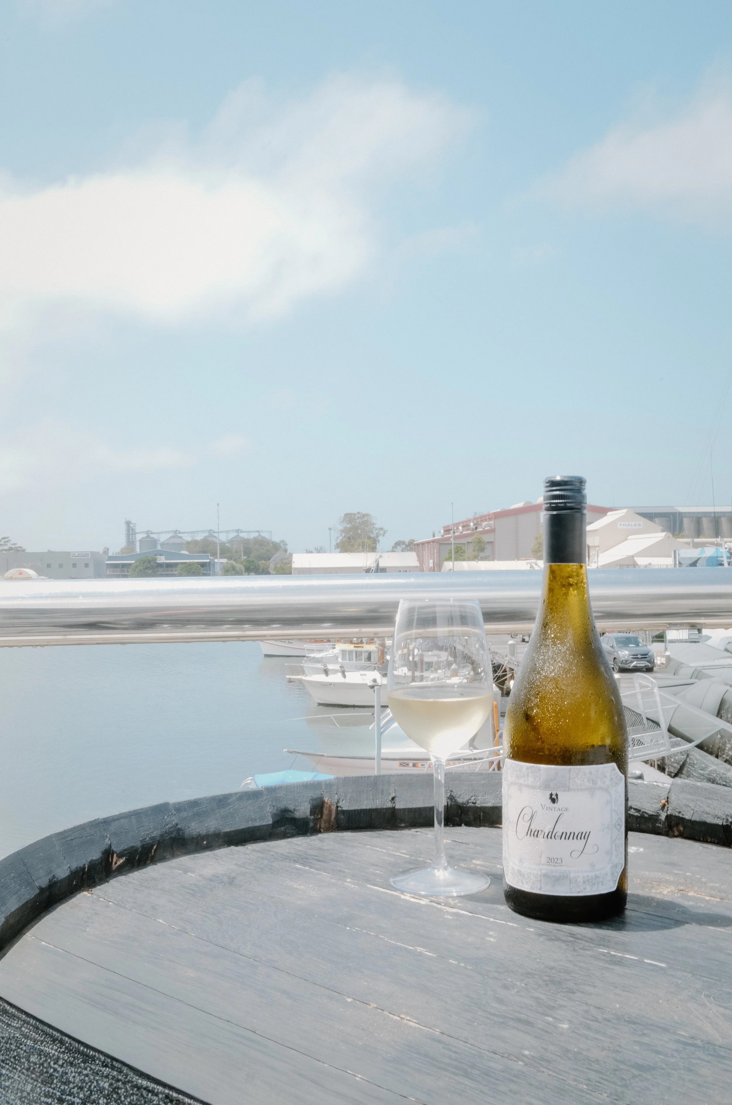 A bottle of Chardonnay and a glass of white wine on a barrel table at a marina with boats and water in the background.