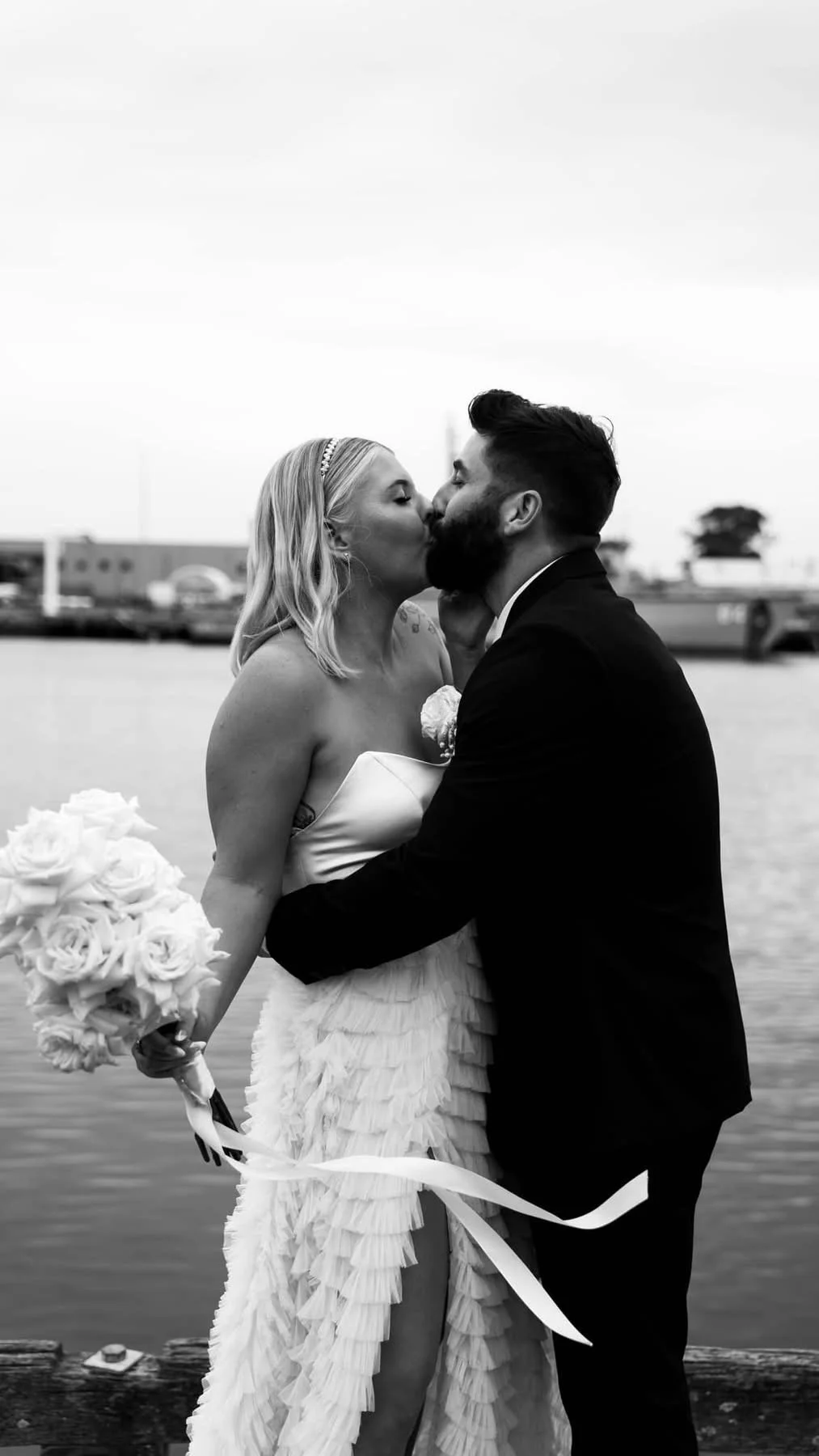 A black and white photo of a bride and groom sharing a kiss on a dock by the water. The bride holds a bouquet of roses, and the groom is dressed in a tuxedo.