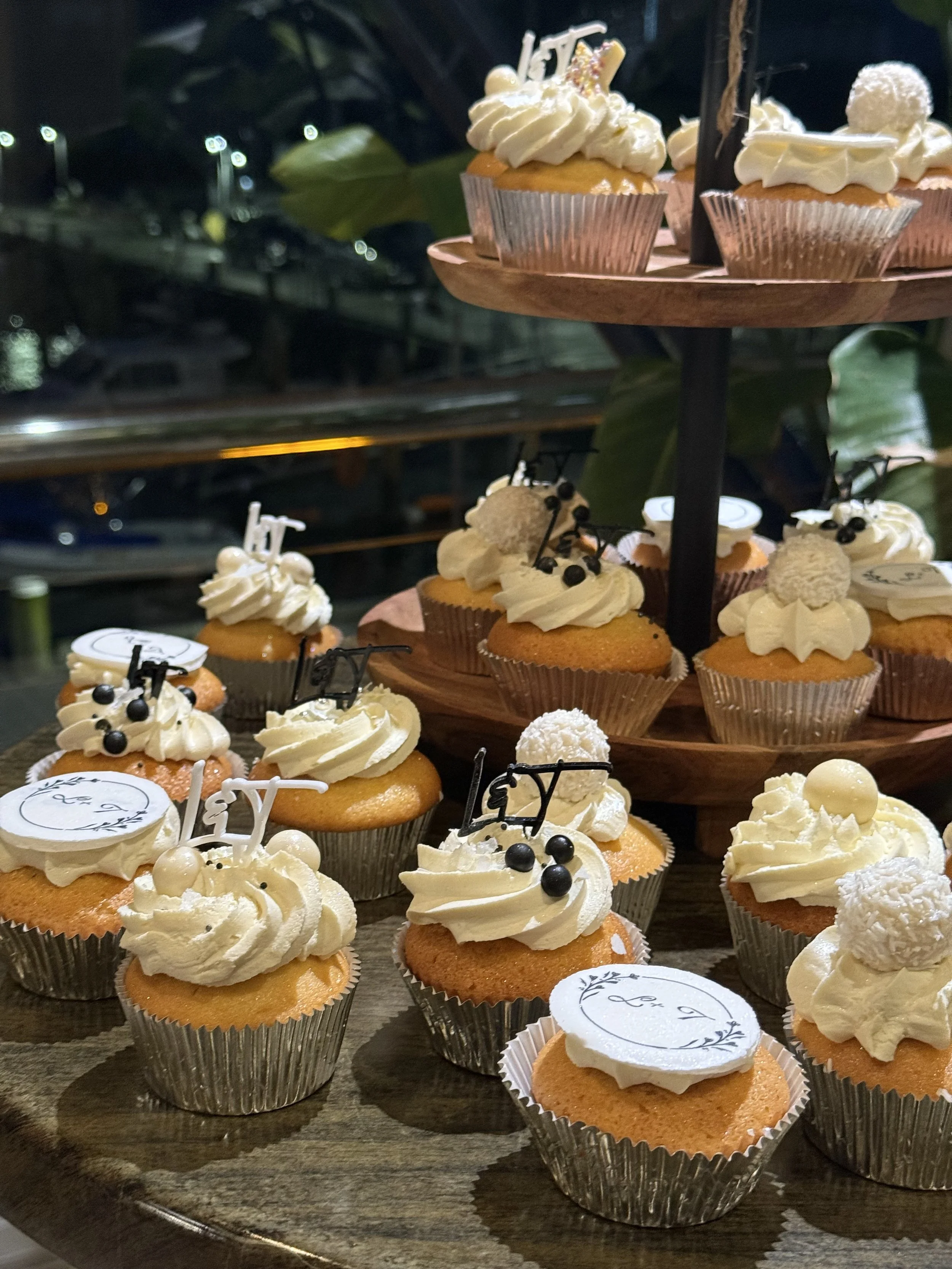 Cupcakes with white frosting, decorative black berries, white chocolate balls, and edible toppers on a multi-tiered wooden display table at night.