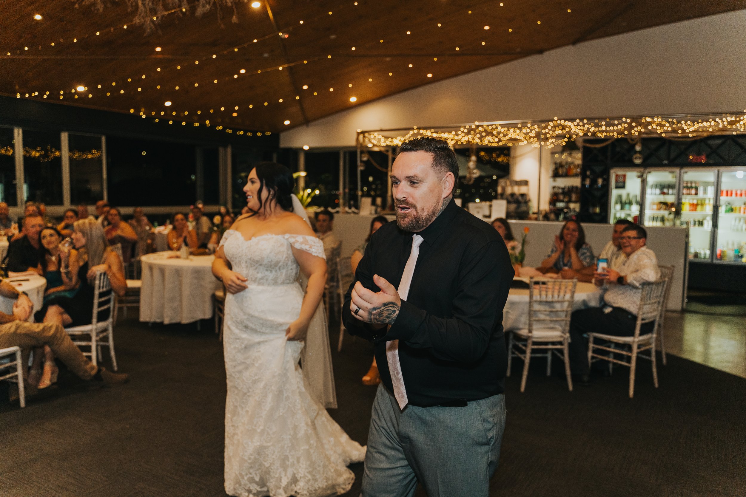 A man and woman dancing at a wedding reception with guests seated around tables in a decorated venue with string lights.
