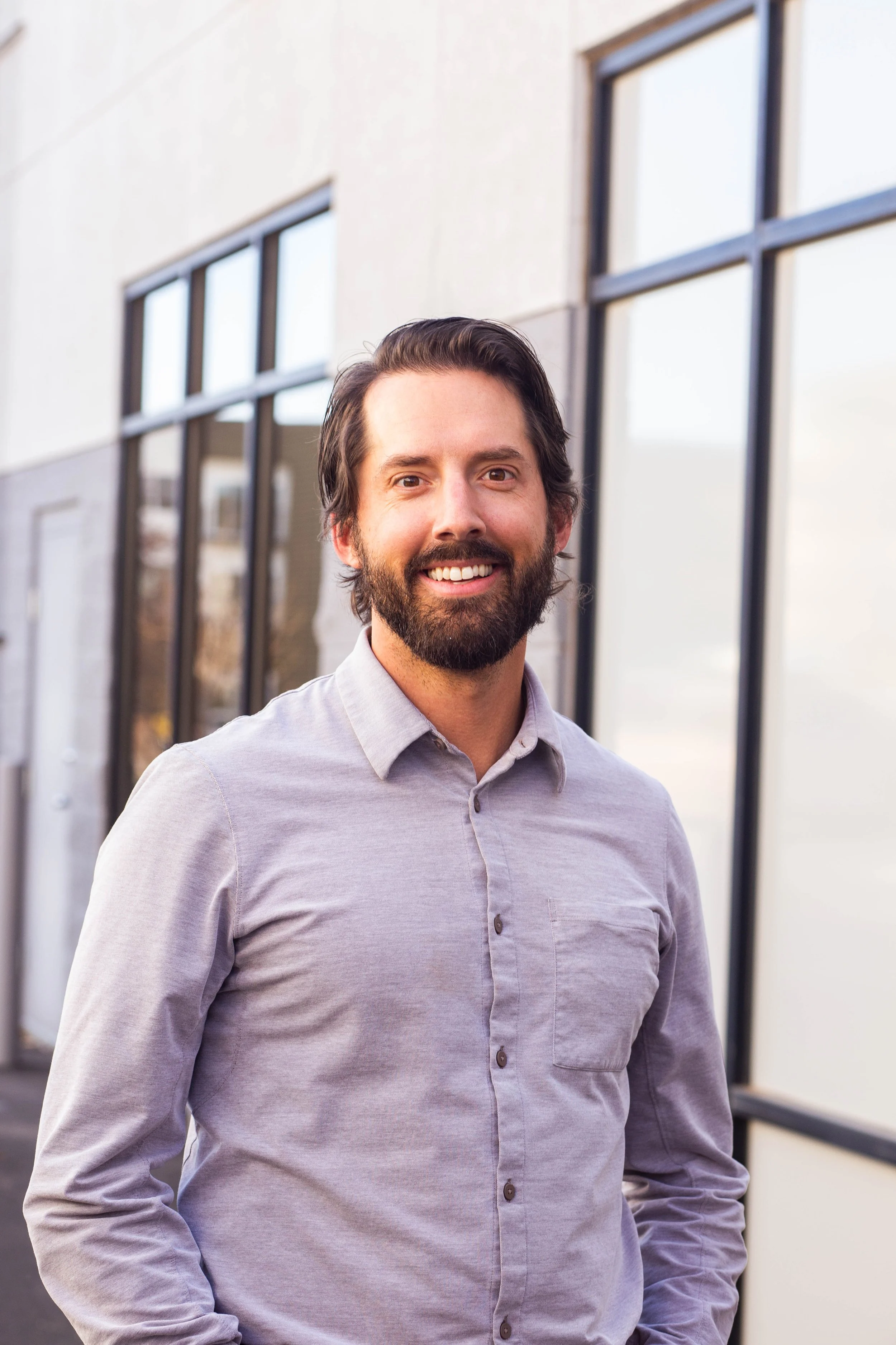 A smiling man with a beard and brown hair standing outside near a building with large windows.