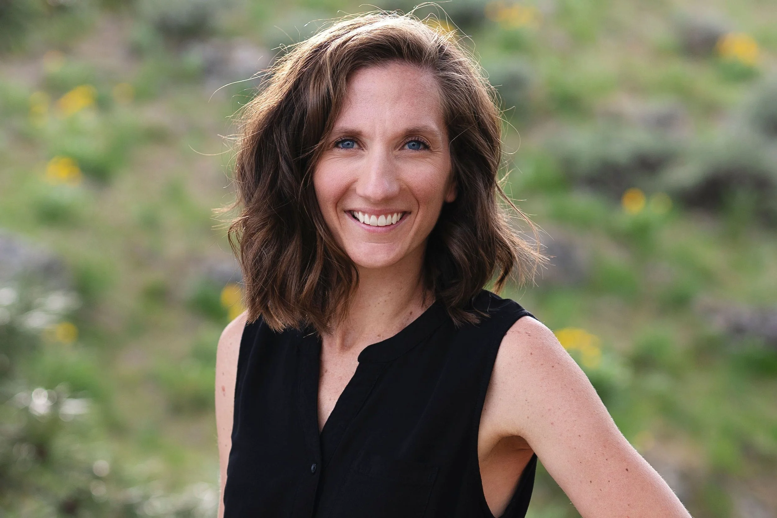 A smiling woman with wavy brown hair wearing a black sleeveless top outdoors with greenery and yellow flowers in the background.