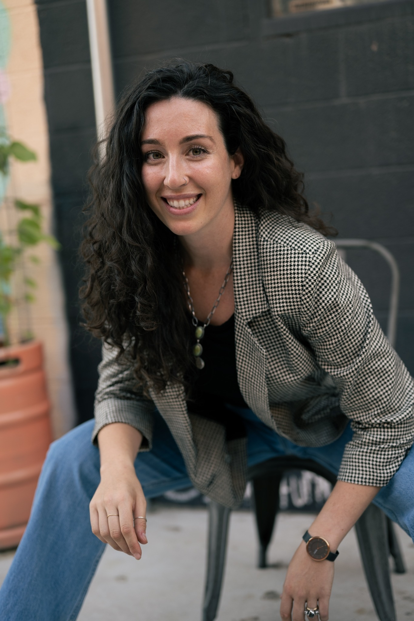Woman with long curly dark hair smiling, sitting outdoors on a black chair, wearing a checked blazer, black top, blue jeans, a necklace, rings, and a watch, in front of a black wall with potted plants nearby.