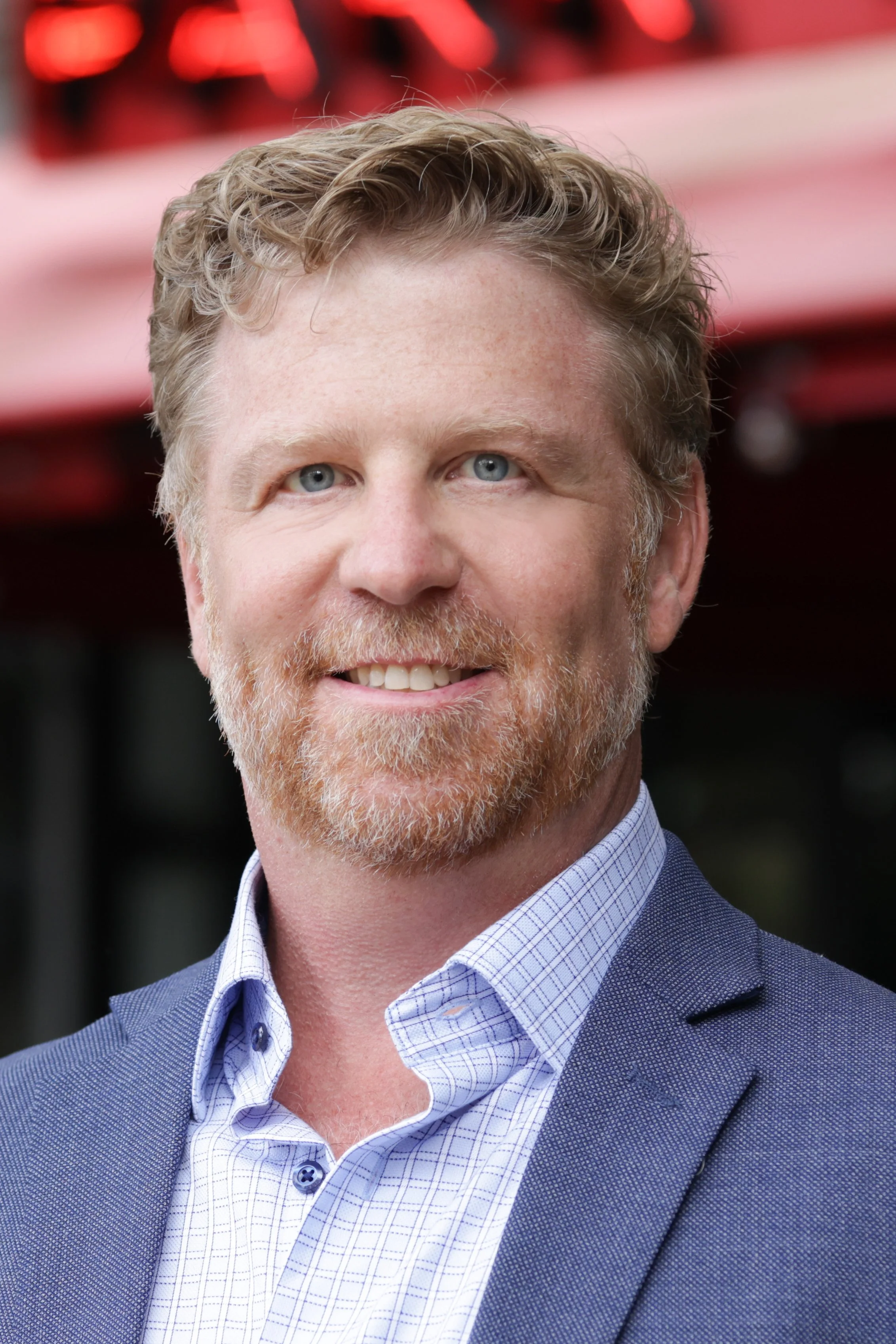A man with light skin, red hair, and a beard, smiling, dressed in a blue blazer and a checkered shirt, standing outdoors in front of a red structure.