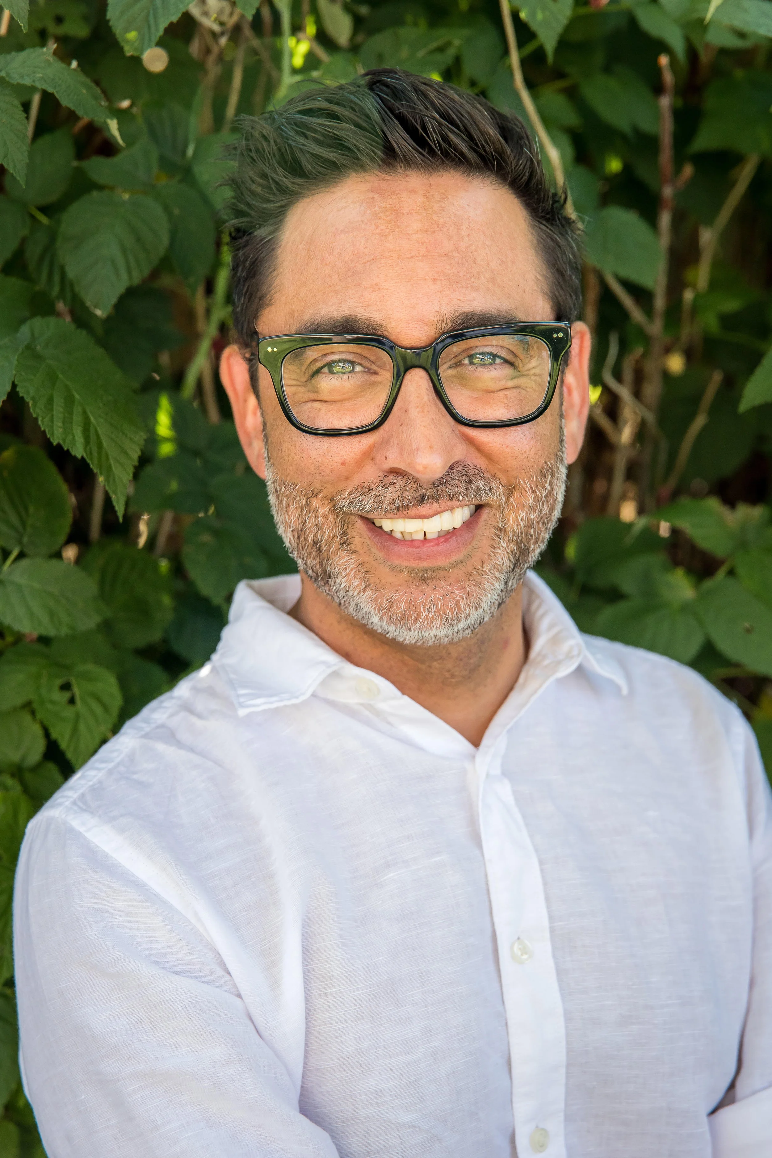 A smiling man with glasses, a beard, dressed in a white shirt, standing in front of green foliage.