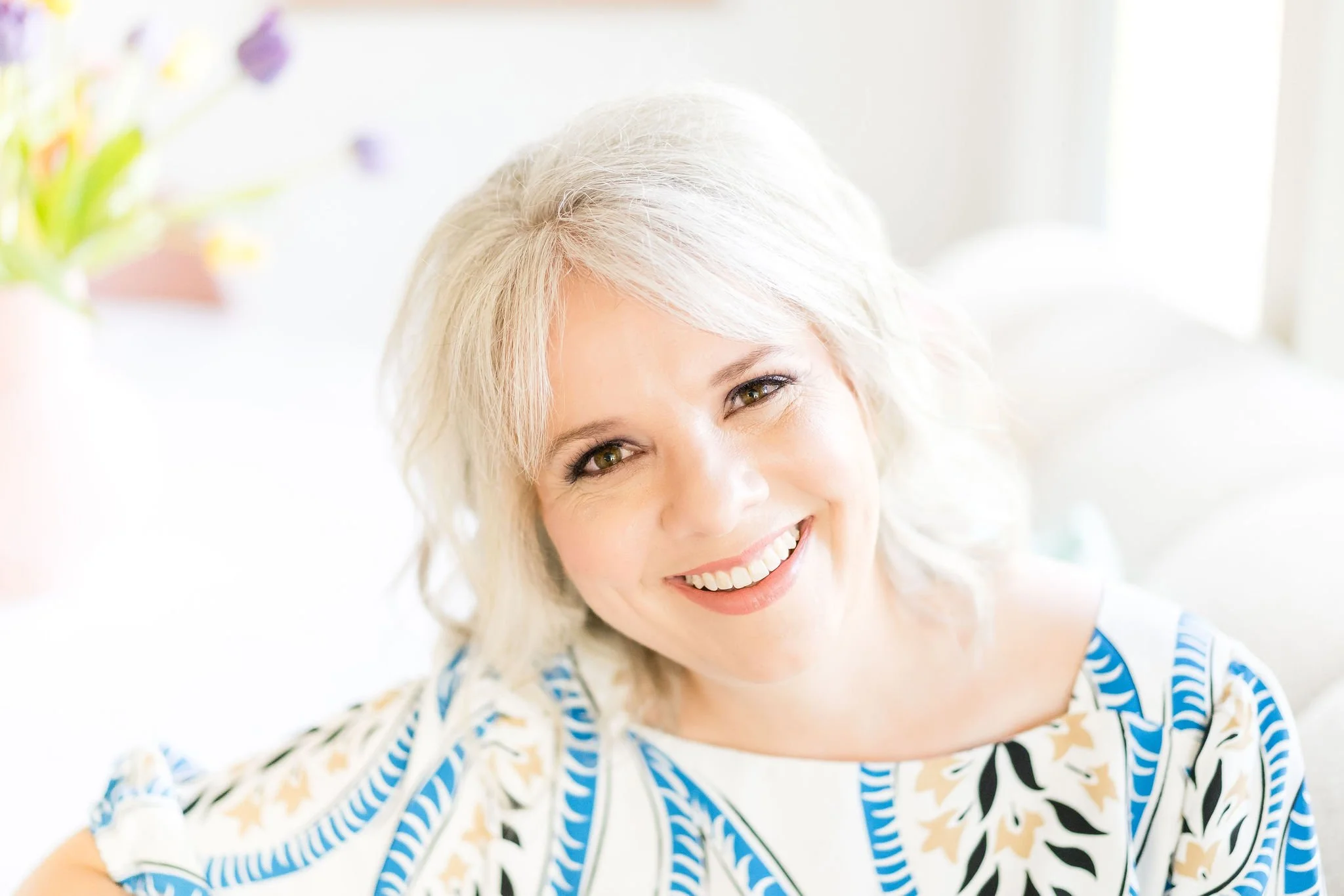A woman with short gray hair, smiling and wearing a green shirt and earrings, resting her chin on her hand outdoors with a blurred water background.