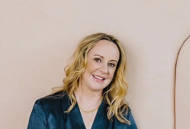 A woman with blonde wavy hair smiling, wearing a dark blue satin blouse and a gold necklace, against a light-colored wall.