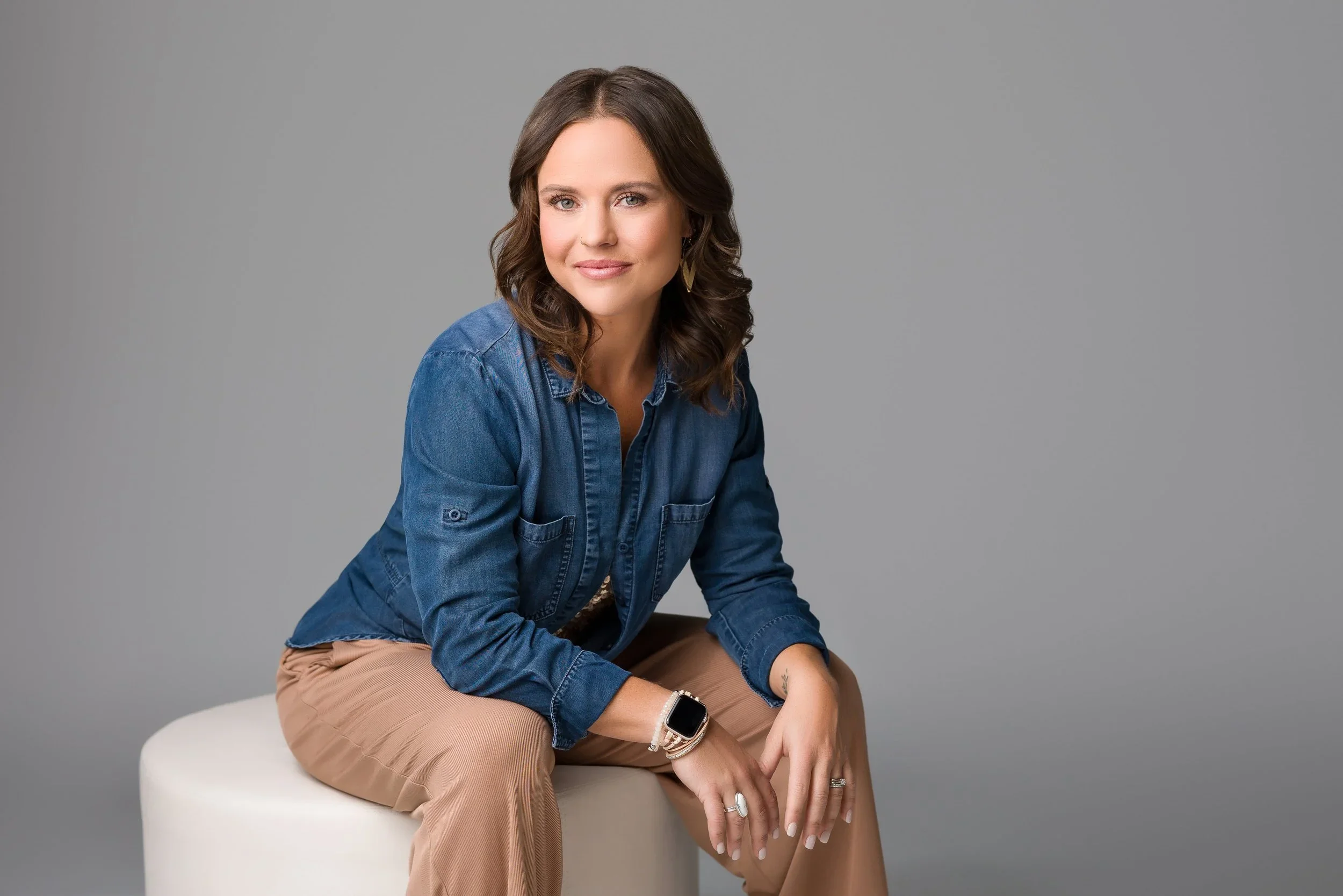 Woman with shoulder-length brown hair, wearing a denim shirt and beige pants, sitting on a white round stool against a plain gray background, smiling at the camera.