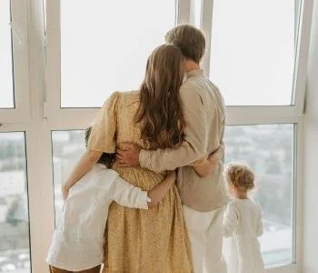 A family of four looking out a large window, with the parents hugging and their two children standing nearby.