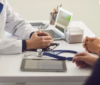 Doctor's office with a doctor and a patient discussing, medical tools on desk, laptop, tablet, and clipboard.