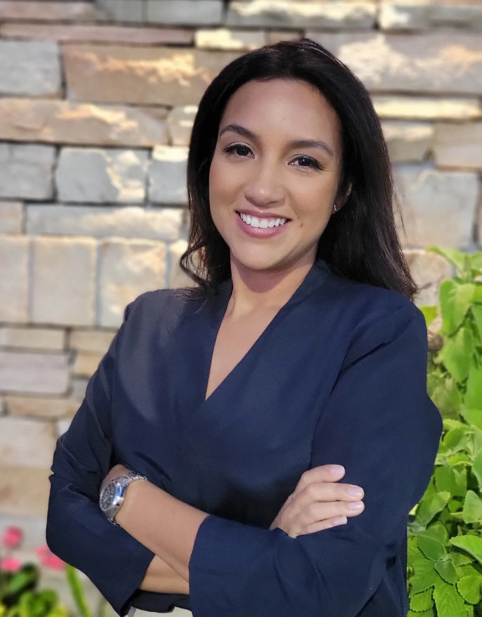 A woman with shoulder-length dark hair, smiling, wearing a navy blue blazer, standing with arms crossed in front of a stone wall and greenery.