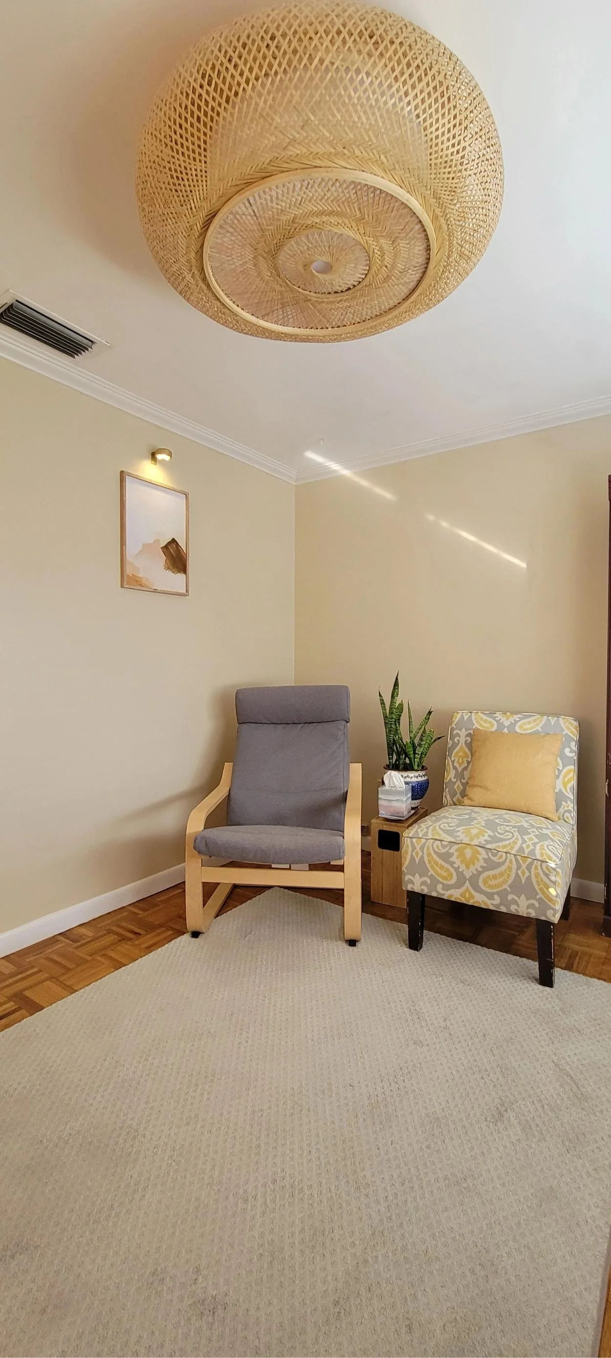 A cozy living room corner with a beige rug, a gray armchair with wooden arms, a patterned accent chair with yellow and gray design, a small wooden side table with tissues and a potted plant, a wall picture of a mountain landscape, and a round woven ceiling light fixture.