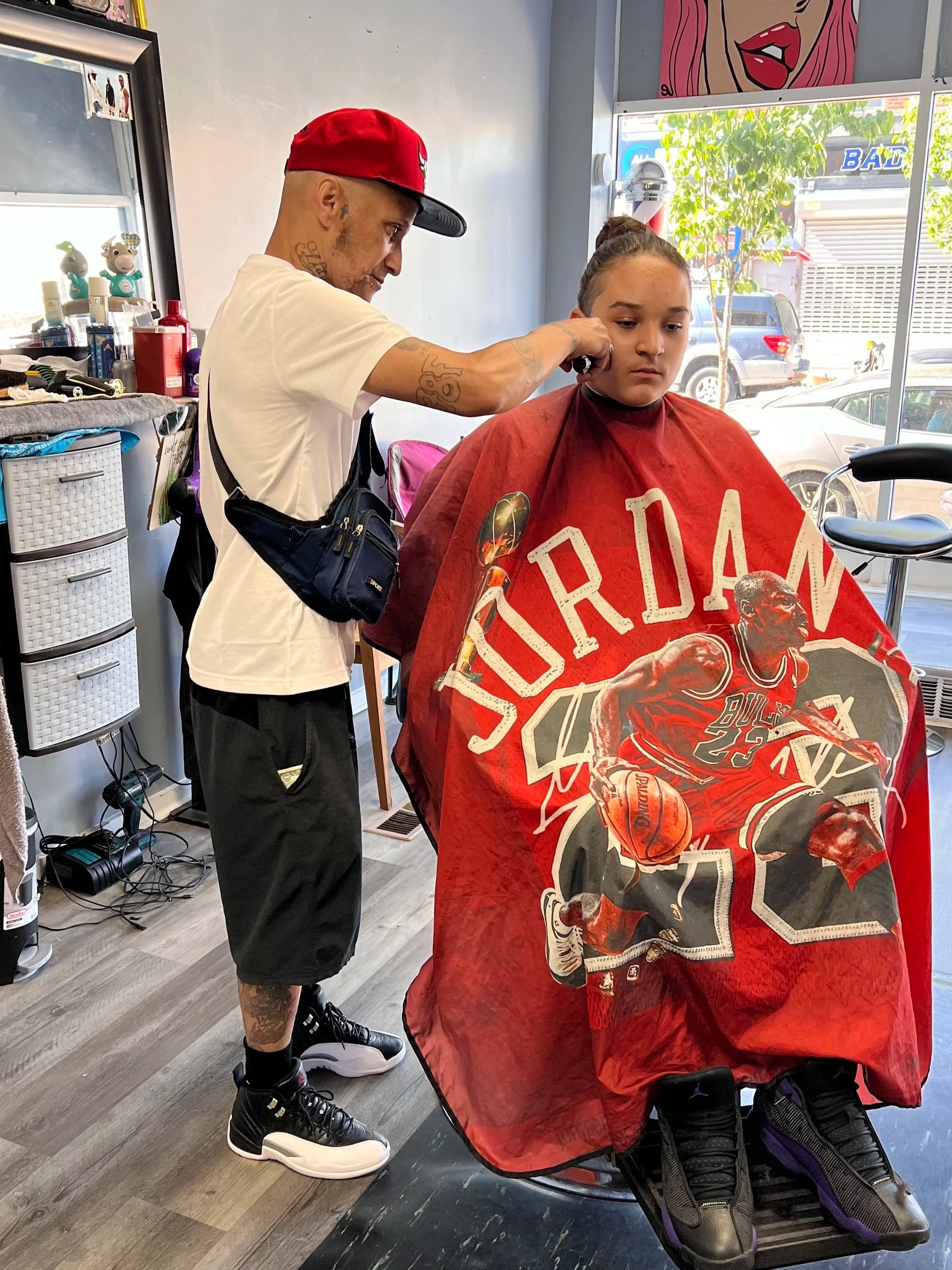 A man with tattoos getting haircut by a barber in a salon, sitting in a haircutting cape with a Michael Jordan basketball design, while the barber trims his hair.