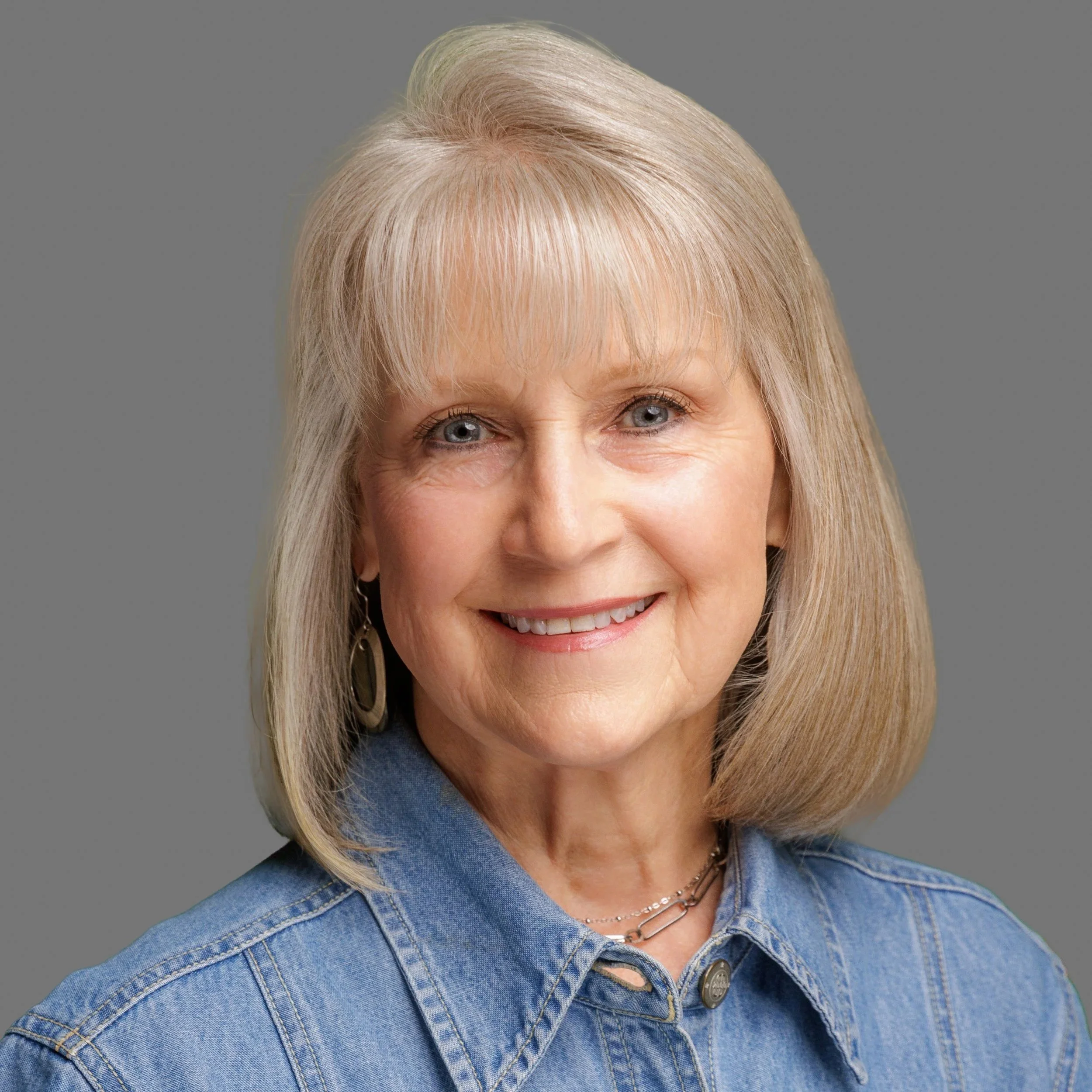 Close-up portrait of a smiling older woman with blonde hair, blue eyes, wearing a denim jacket, earrings, and a necklace, against a plain gray background.