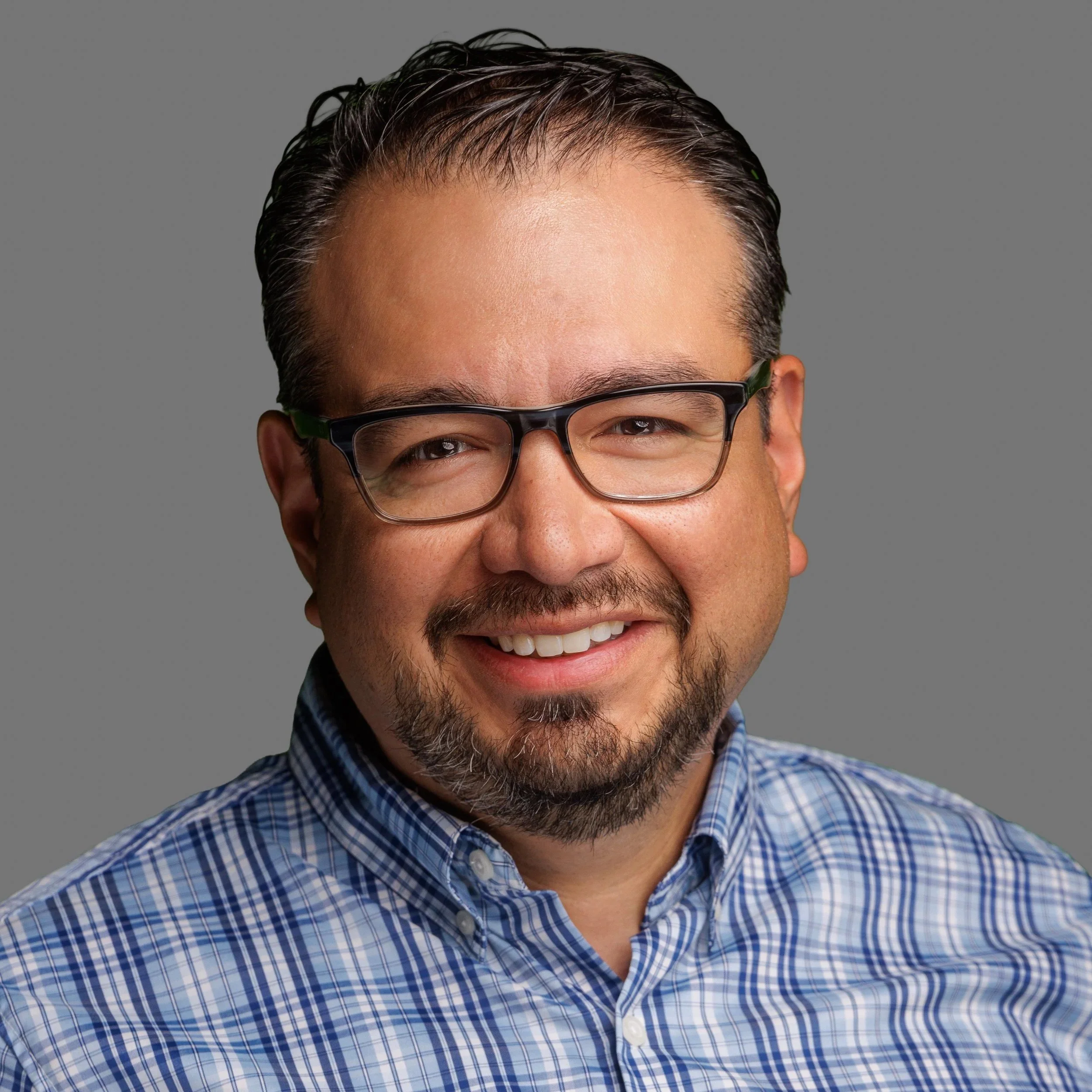 A smiling man with dark hair, a goatee, and wearing a blue collared shirt, in front of a wooden background.