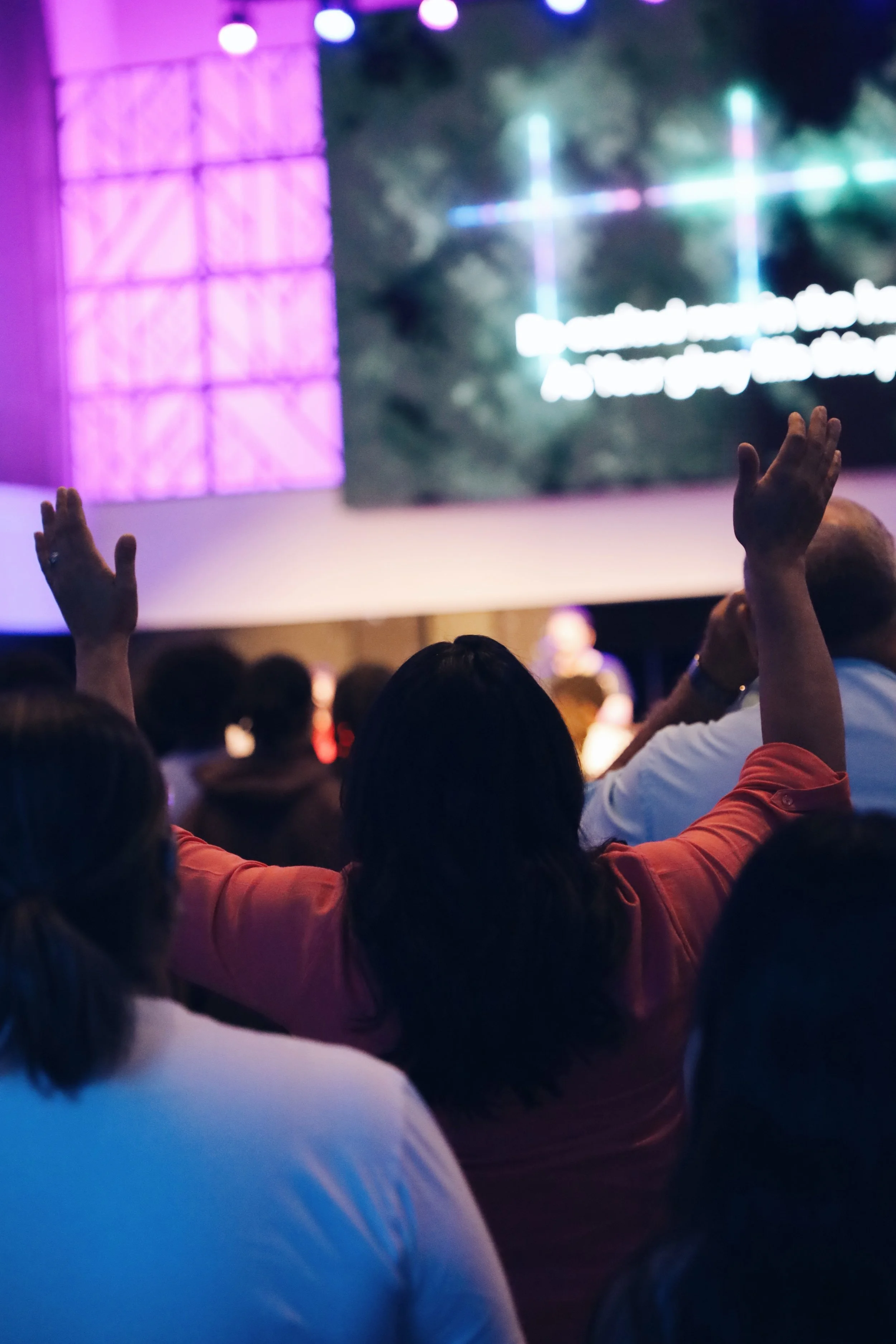 A person in a reddish shirt raising their hand in an audience at a conference or presentation, with a large screen in the background displaying colorful graphics and text.