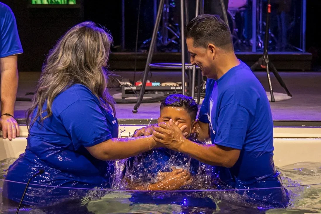 A child baptizing in a baptism ceremony with two adults helping in a pool, water splashing around, stage with musical equipment in the background.
