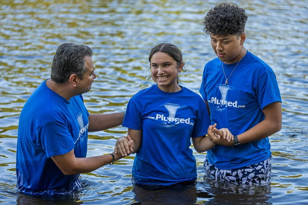 A group of three people standing in a body of water, smiling and holding hands. They are wearing matching blue T-shirts with the logo 'Plunged.me' on them.