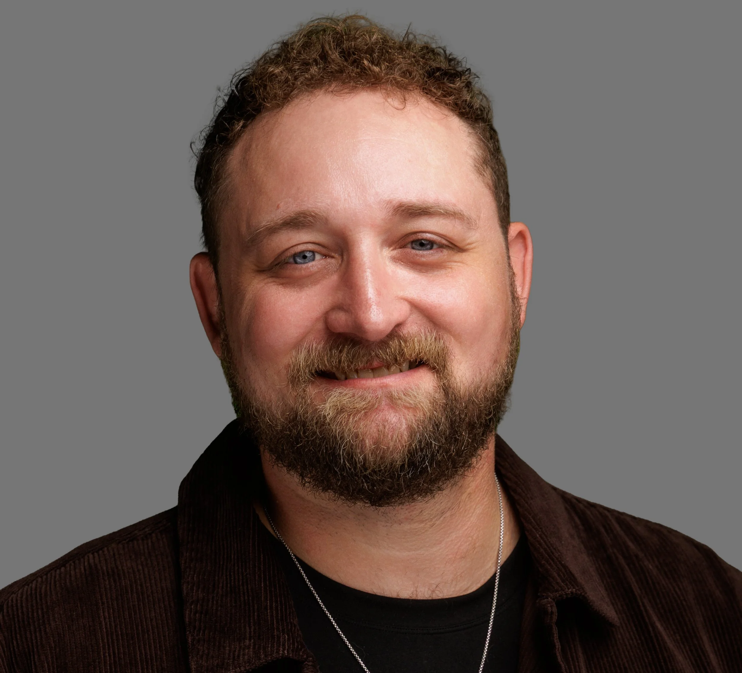 A man with light skin, a beard, and blue eyes, wearing a brown shirt, smiling at the camera against a grey background.