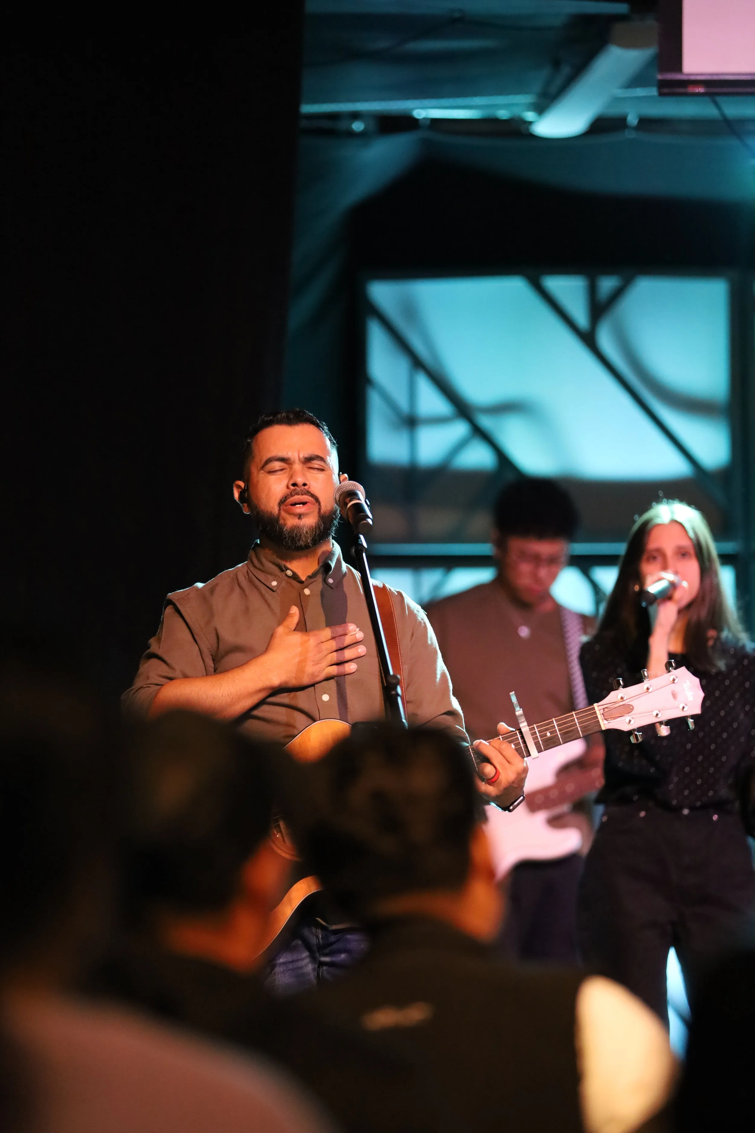 A man singing with a guitar and a hand on his chest, a woman singing with a microphone, and another man playing guitar on stage, with an audience in front.