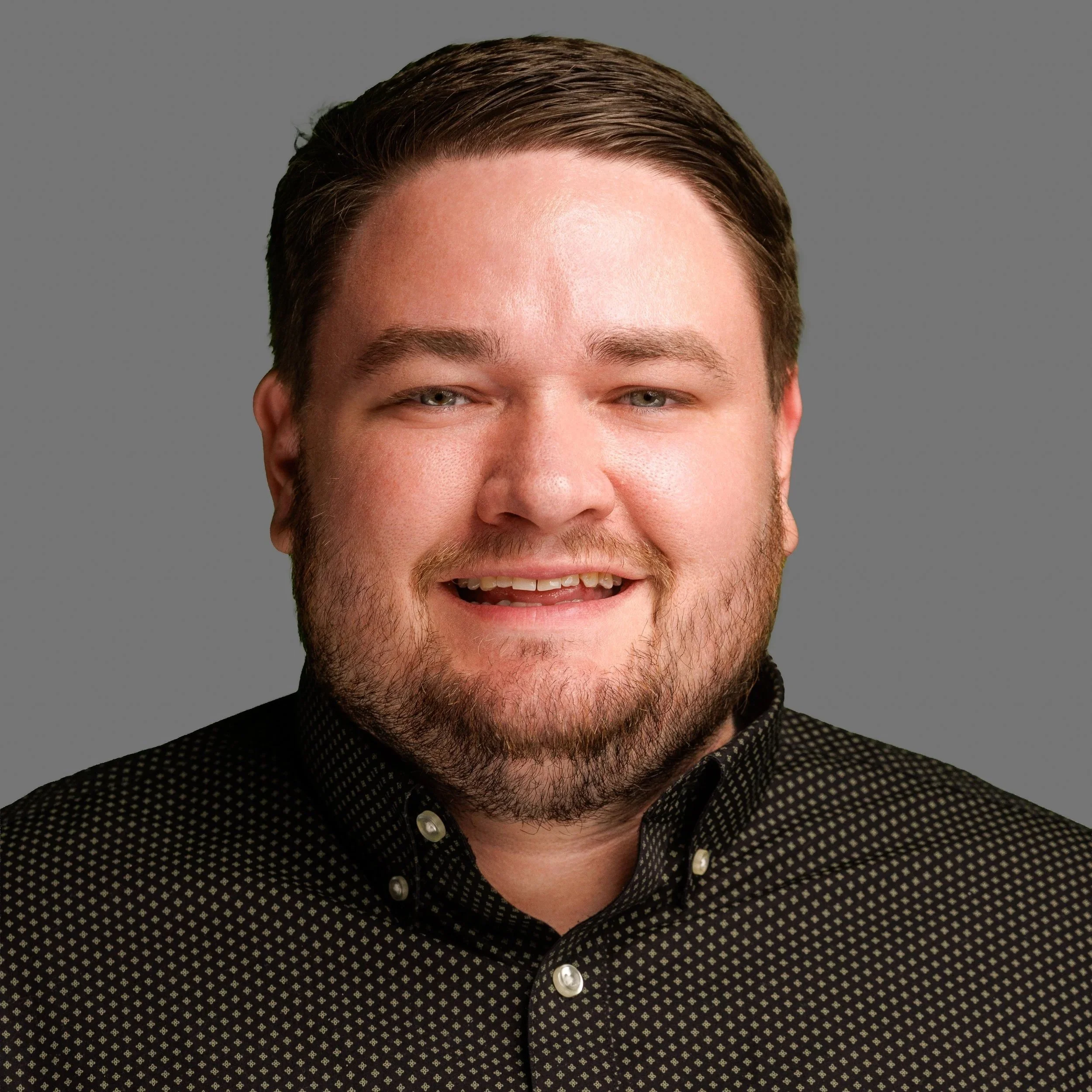 A smiling man with short brown hair, a beard, and mustache, wearing a green collared shirt, standing indoors against a gray background.