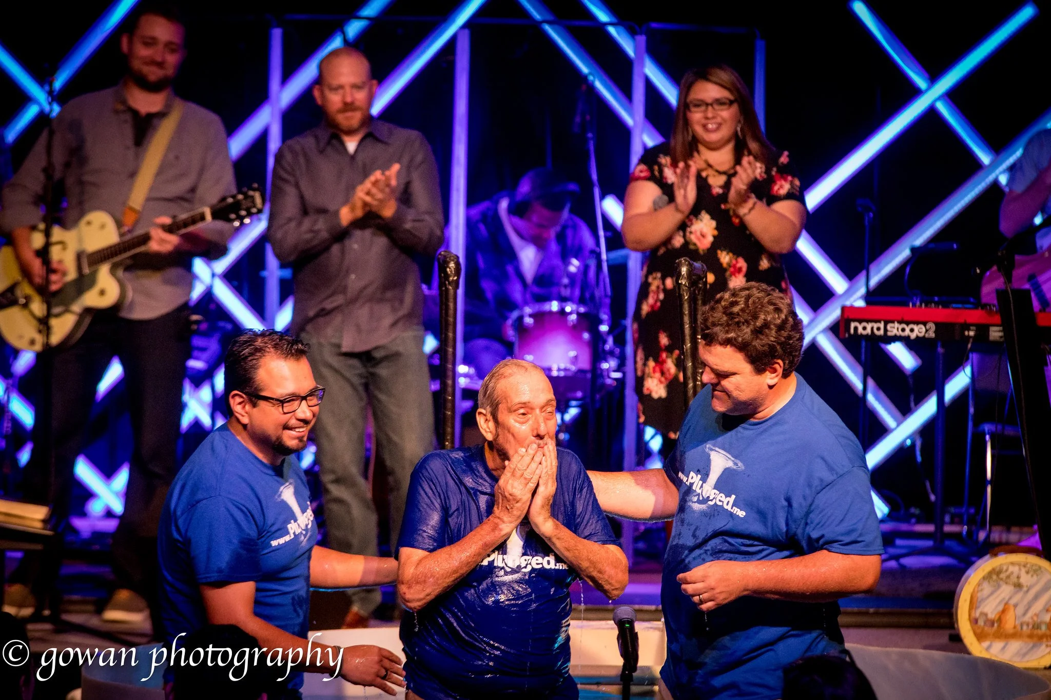 An older man coming out of the water being supported by two men in blue shirts during a church baptism ceremony, with a band playing in the background on stage.