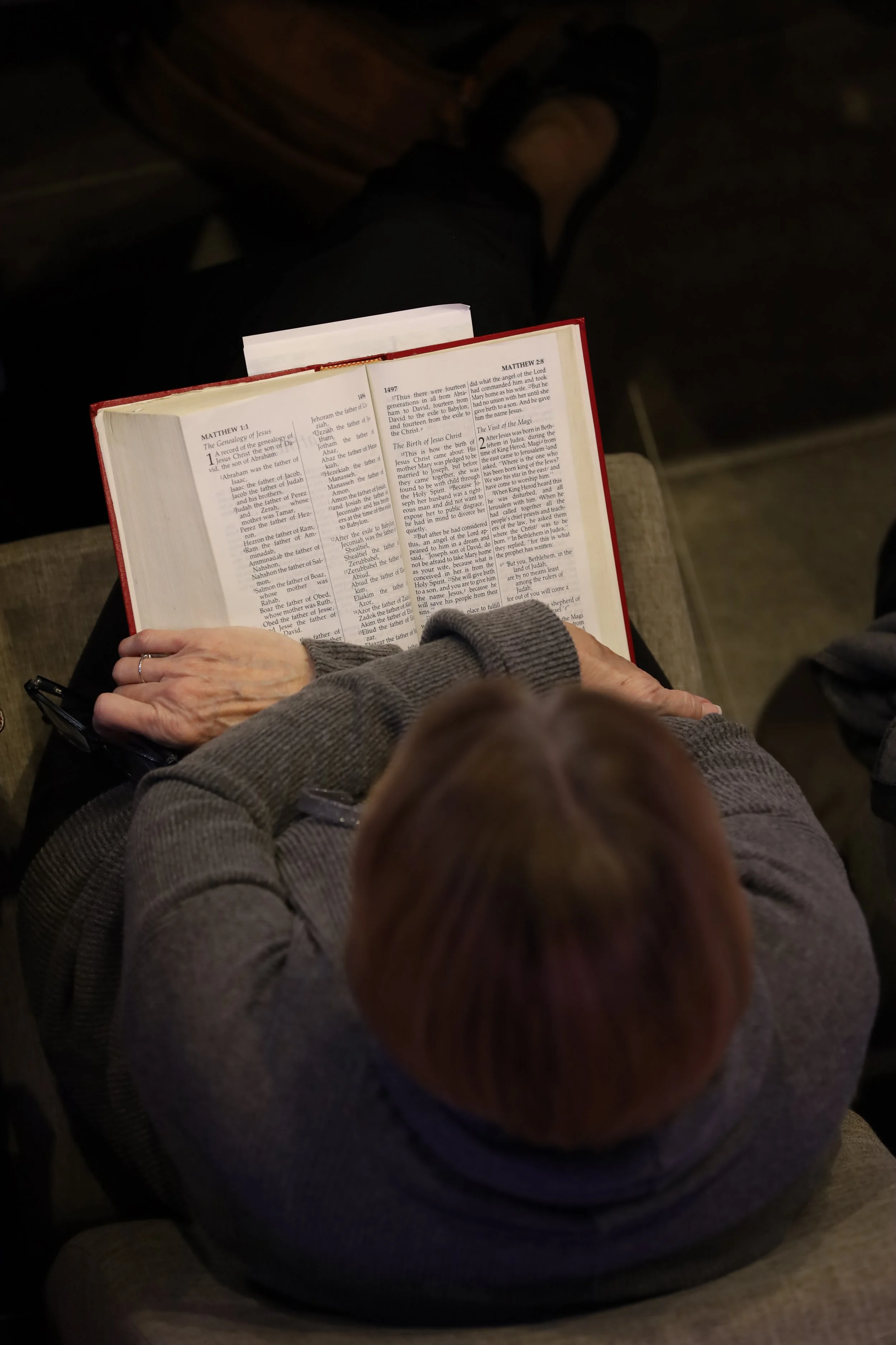 A person with short, brown hair is sitting in chur, reading a large open Bible with red cover. The person is wearing a gray sweater and glasses resting on their lap. The focus is on the Bible from above, with legs and shoes visible in the background.