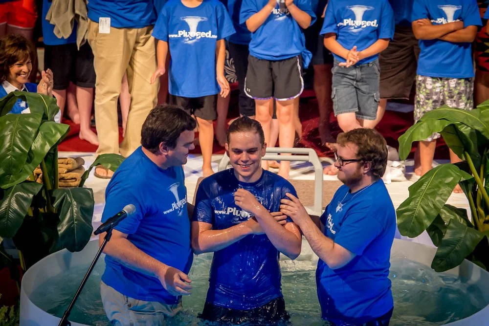 Young man in baptismal water being baptized by two men, surrounded by people in matching blue shirts.