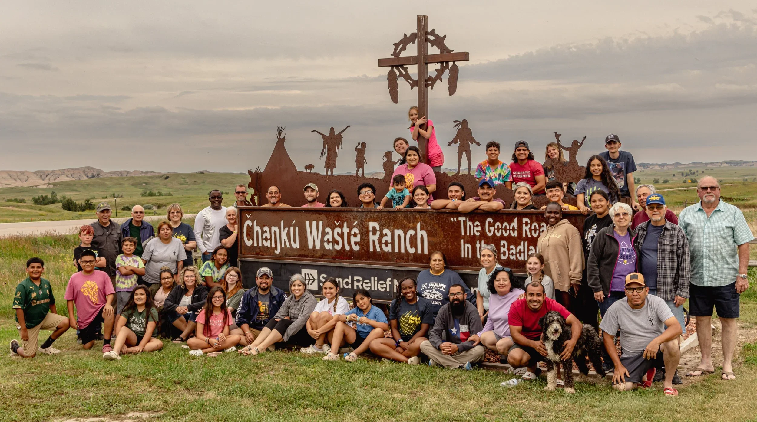 A large group of people gathered outdoors in front of a rust-colored sign that reads "Chanku Waste Ranch. The Good Road in the Badlands." The group includes children and adults, some seated on the grass and others standing behind the sign. In the background are open fields and rolling hills under a cloudy sky.
