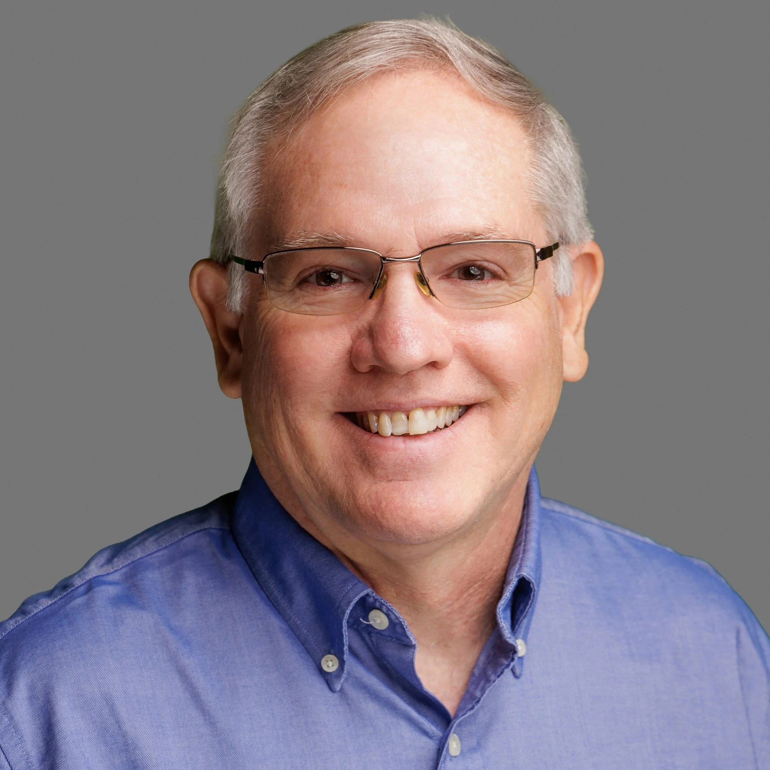 Portrait of a middle-aged man with gray hair, glasses, smiling, wearing a blue collared shirt, against a gray background.