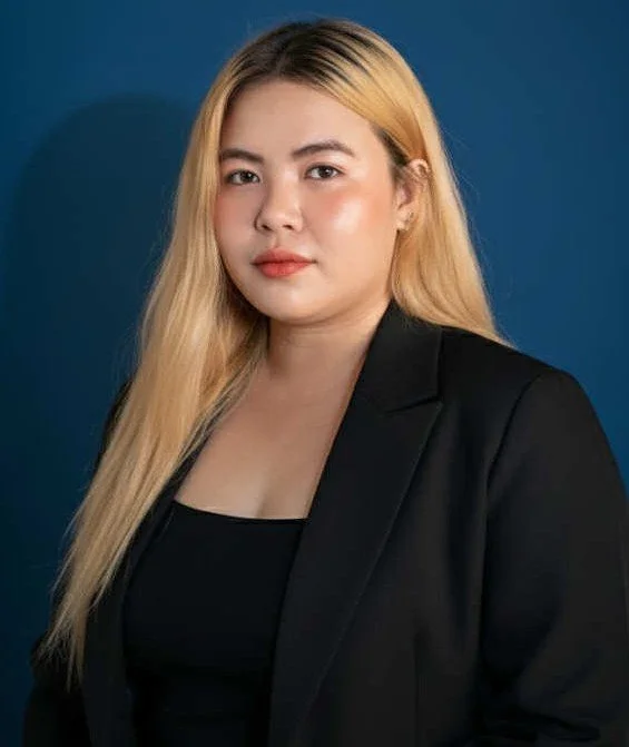 A woman with long blonde hair wearing a black blazer and top, standing against a dark blue background.