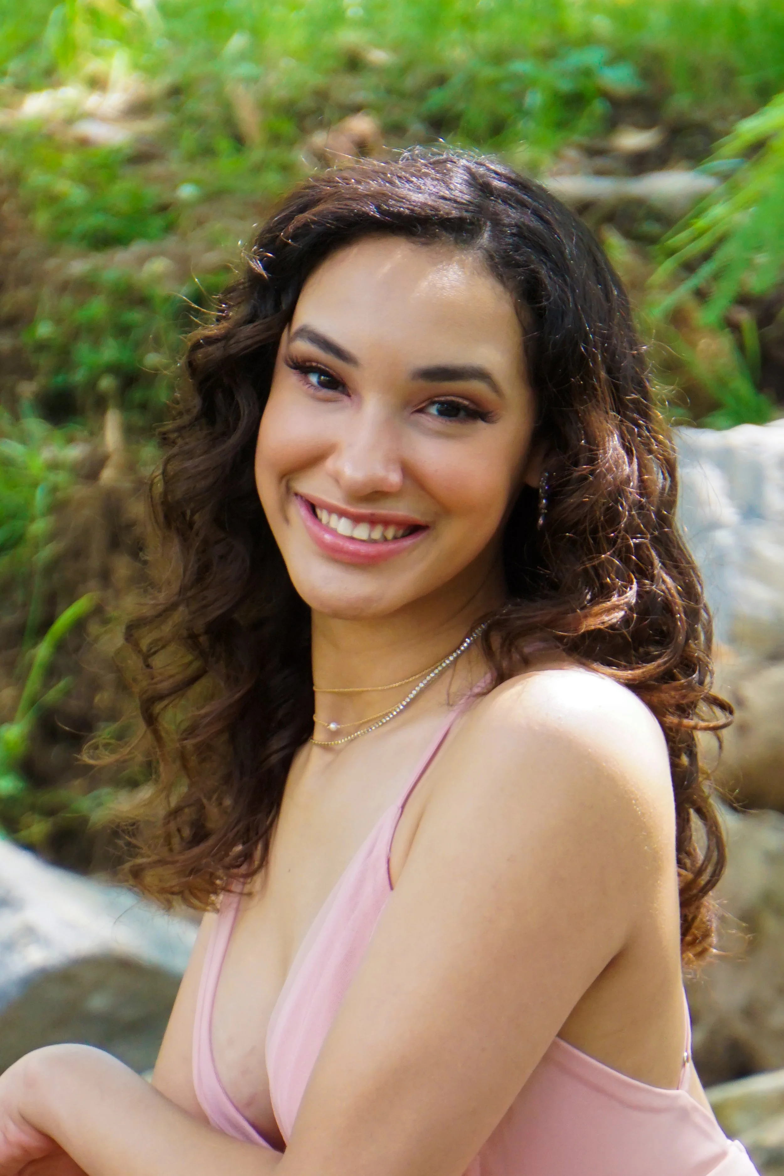 A young woman with brown curly hair smiling outdoors near green foliage and rocks.